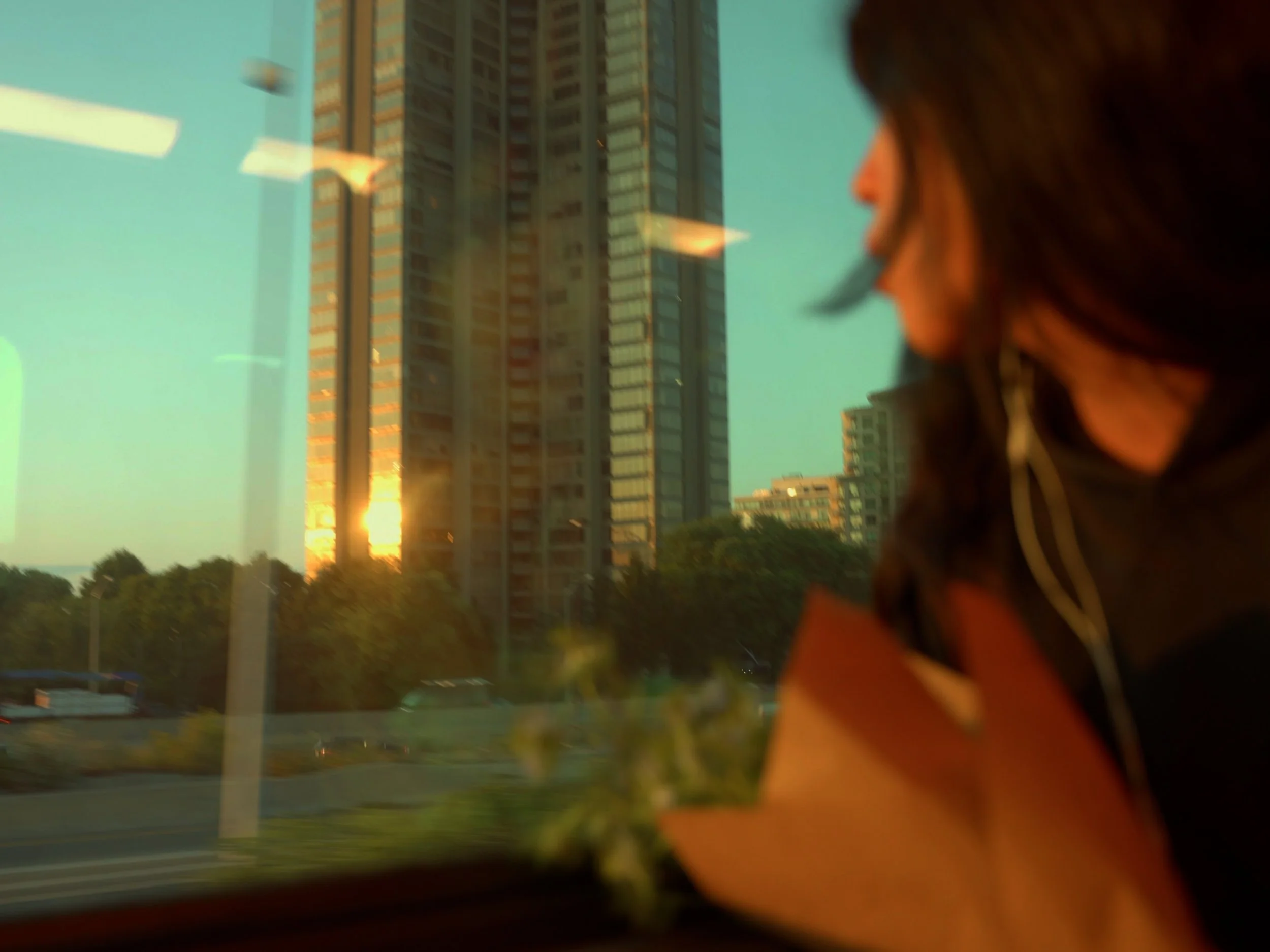 A woman with dark hair looking out a window at tall buildings during sunset.