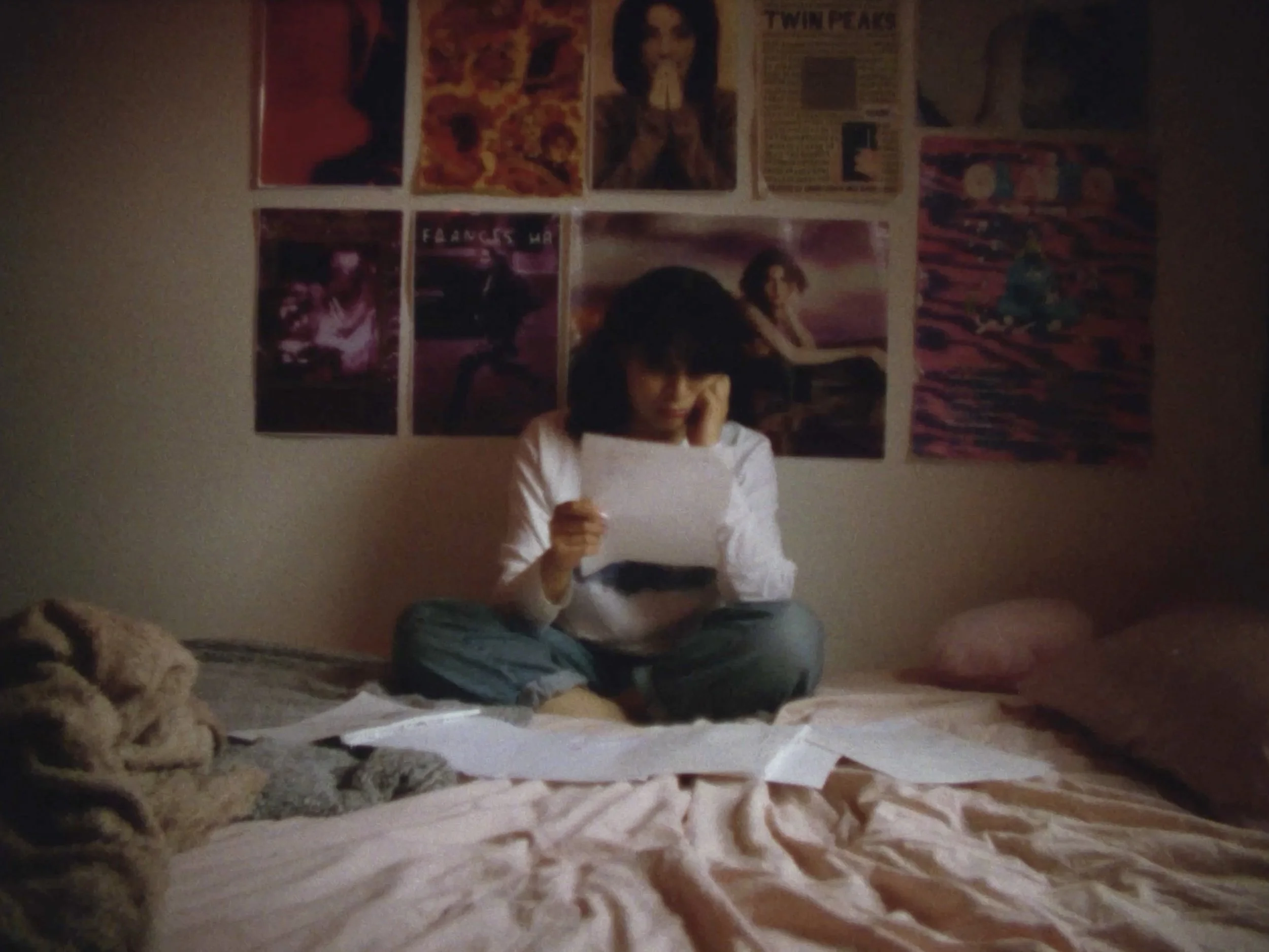 A woman with dark hair sitting on a bed covered with papers, wearing a white long-sleeve shirt and green pants, holding a sheet on paper up. Posters cover the wall behind her.