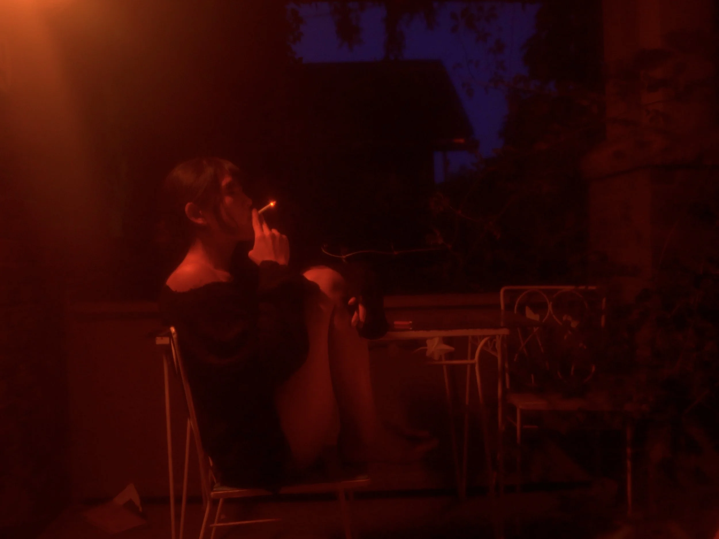 A woman smoking a cigarette in a dimly lit, reddish environment, with some structures or furniture visible in the background.