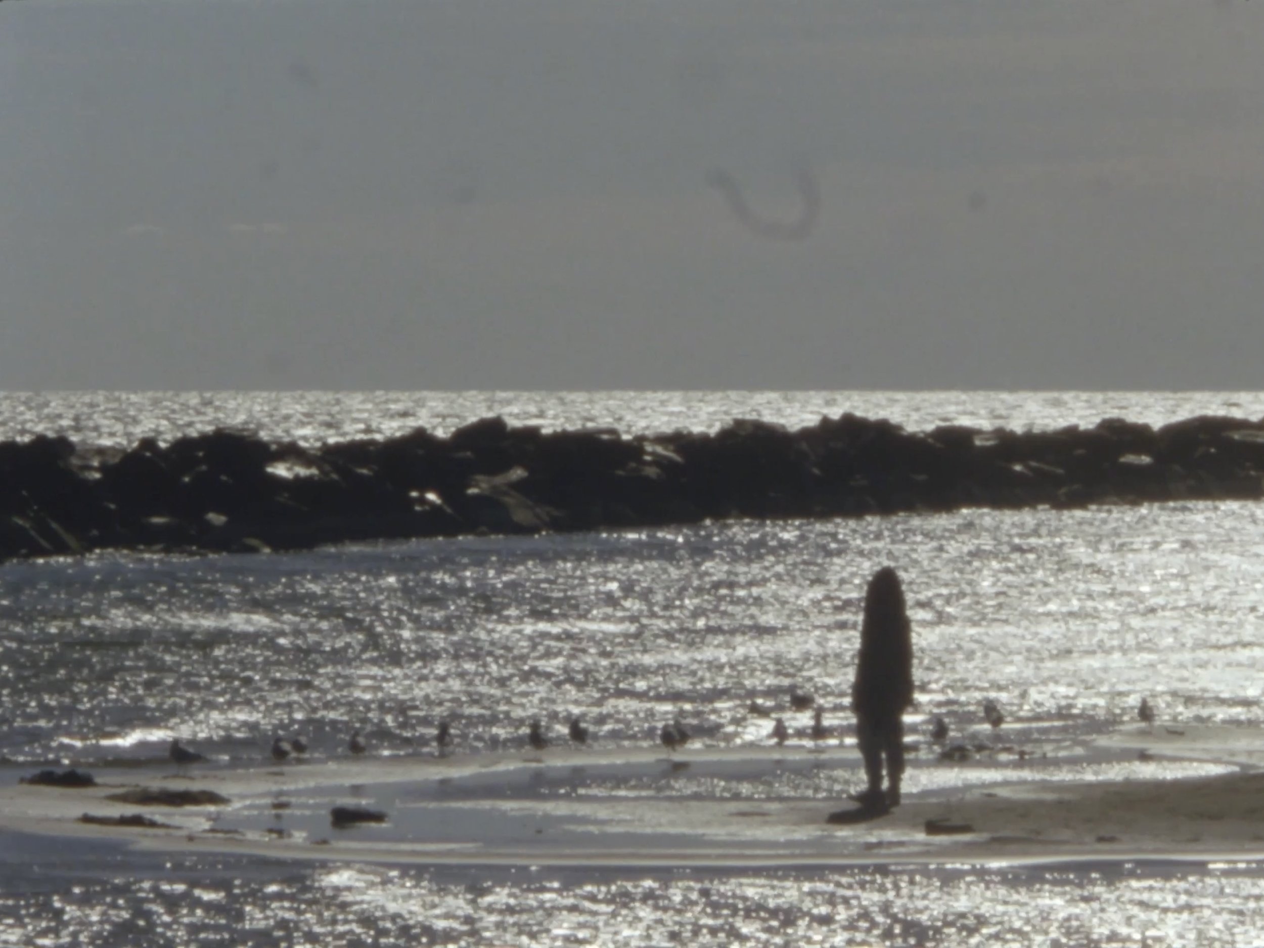 A shadow-y figure standing on a beach at sunset, looking across the water with the sun reflecting off the ocean.