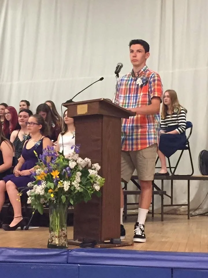 A young man in a colorful plaid shirt and khaki shorts stands at a podium, speaking into a microphone during a ceremony, with a group of young students seated behind him.