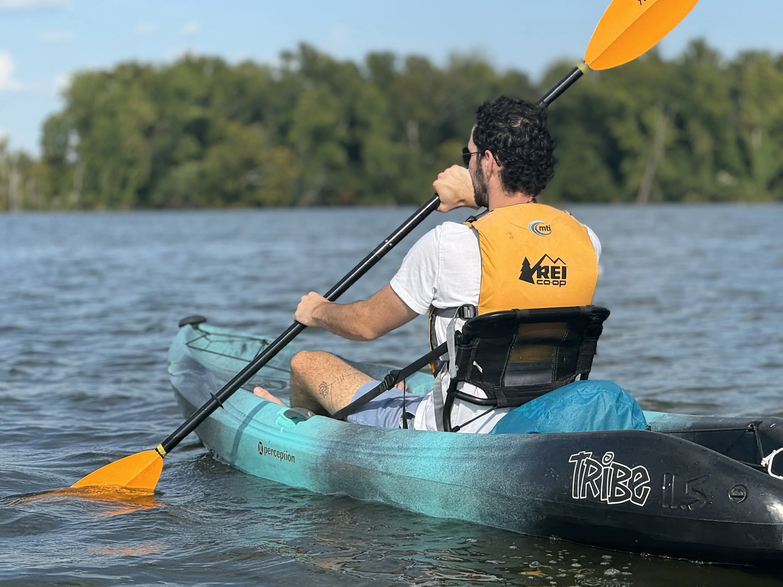 Man kayaking on a lake with green trees in the background, wearing sunglasses and a yellow life vest.