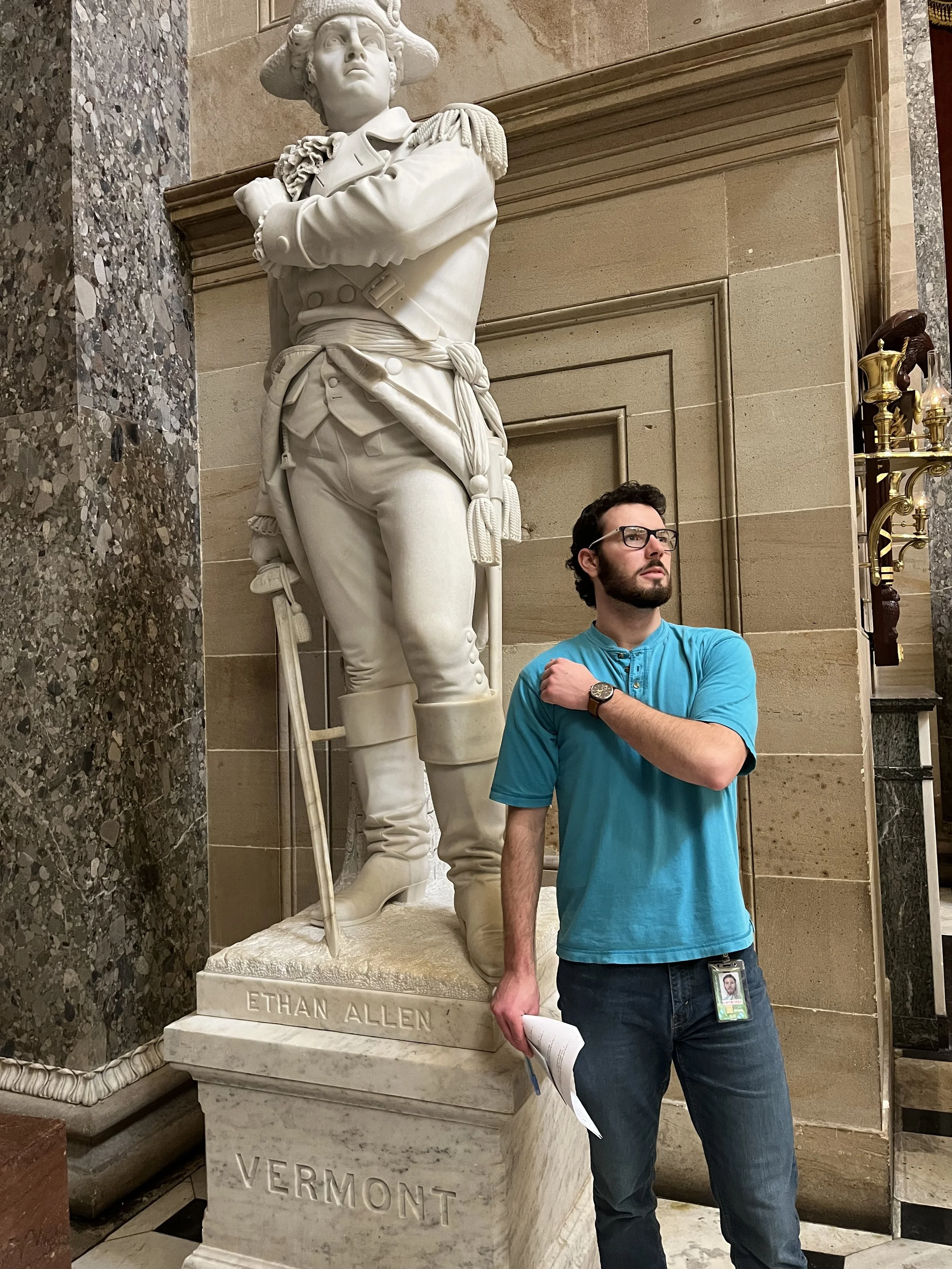A man with glasses, a beard, wearing a turquoise shirt and jeans standing next to a white marble statue of Ethan Allen, holding a rolled-up paper in his right hand inside the US Capitol.