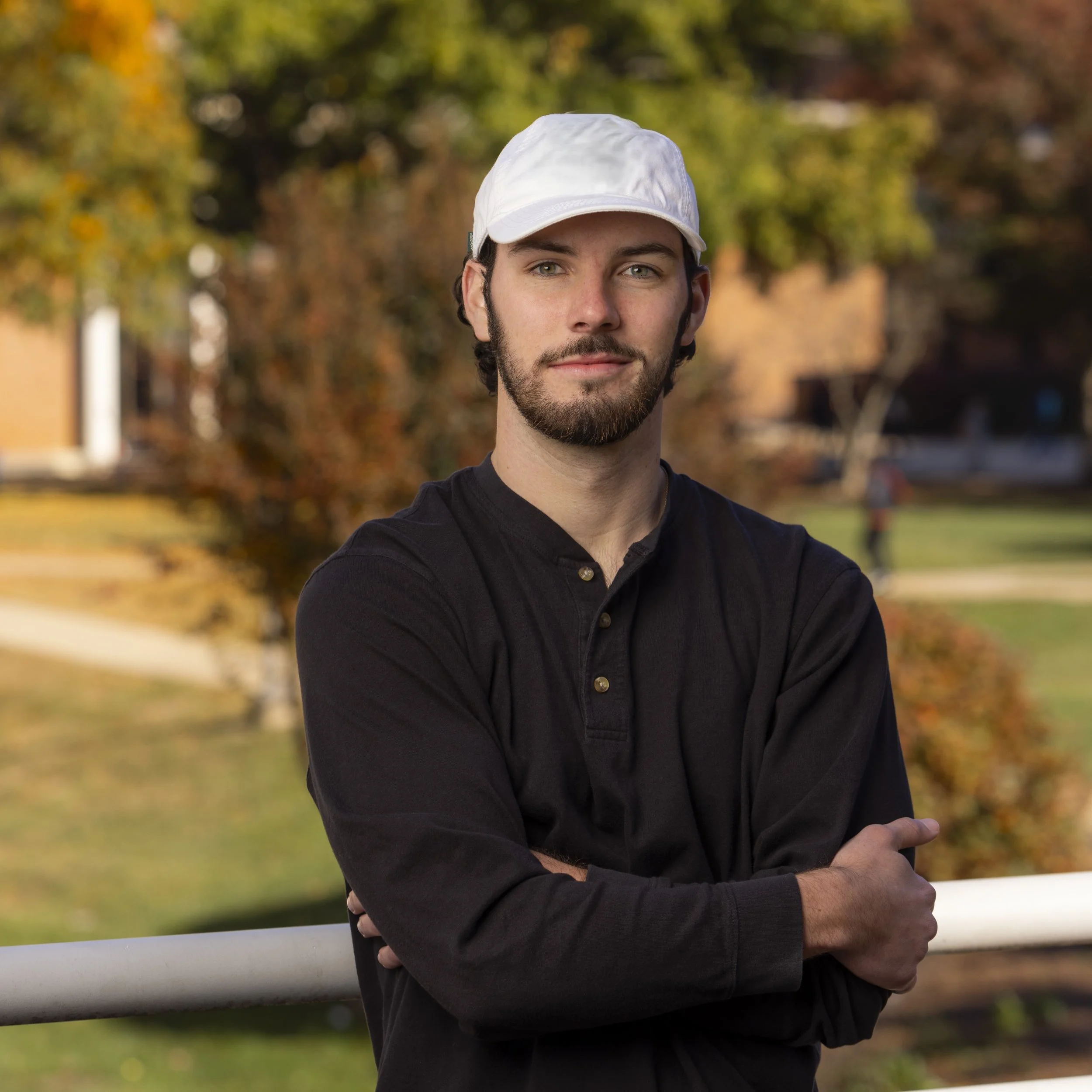 A young man with light skin, dark hair, and a beard wearing a white cap and a black long-sleeve shirt standing outdoors with trees and buildings in the background during autumn.