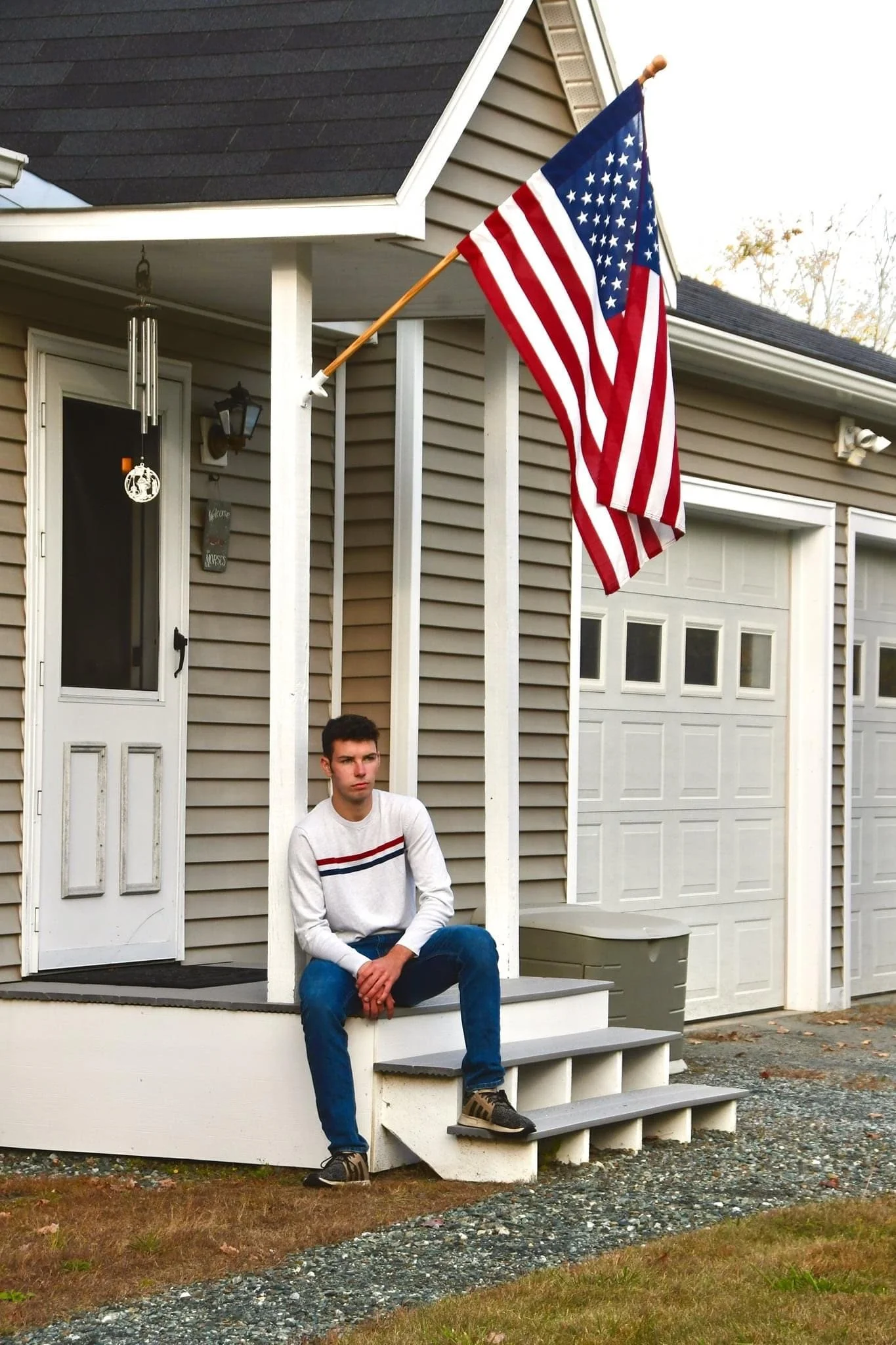 A young man with dark hair sitting on stairs in front of a house, with an American flag hanging from a pole attached to the house.