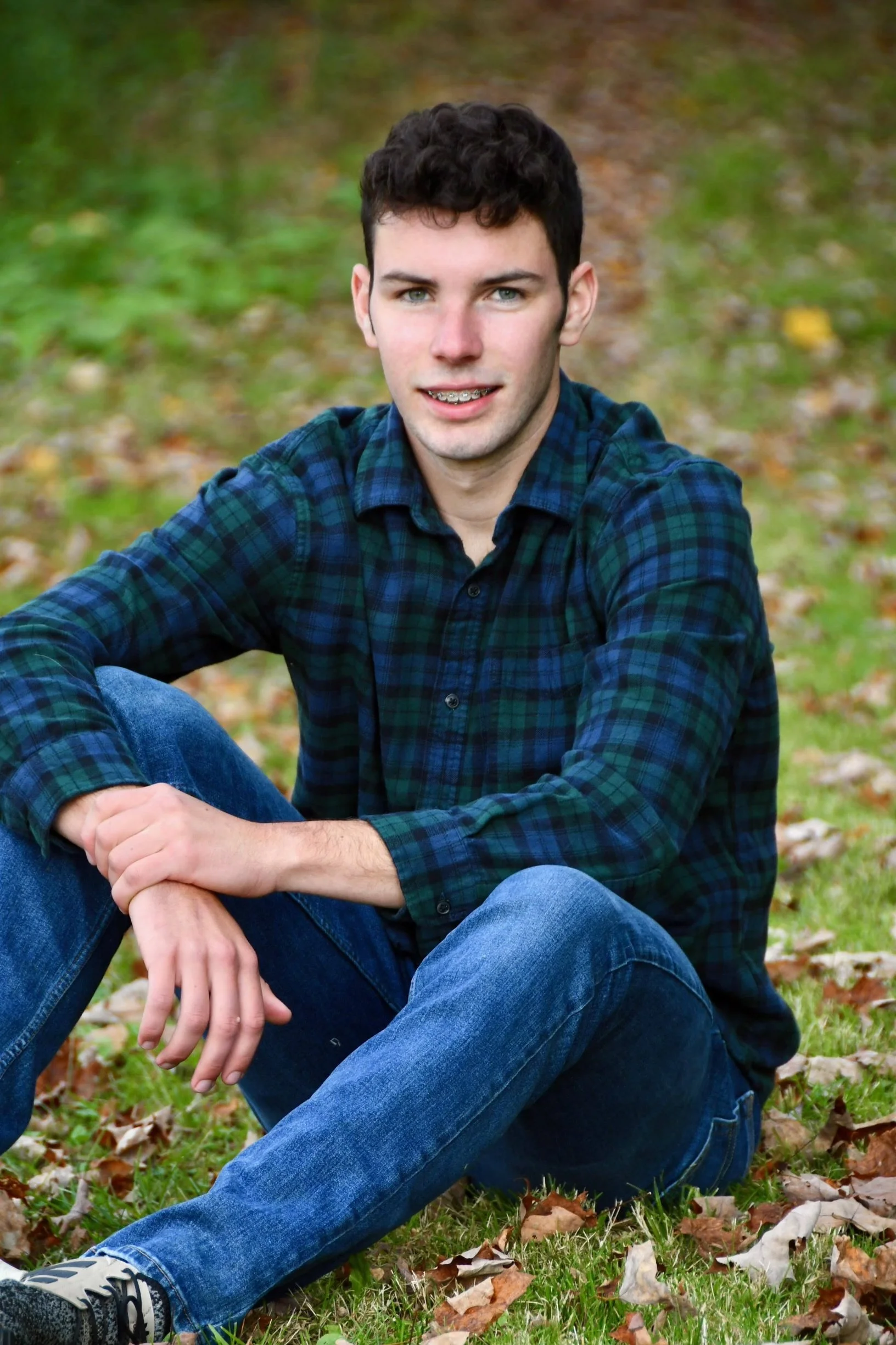 A young man with dark, curly hair and blue eyes sits on the grass surrounded by fallen leaves, wearing a blue and green plaid shirt and blue jeans, looking directly at the camera.