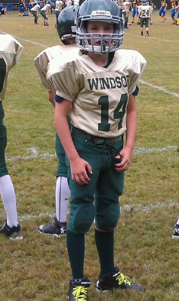 A young football player wearing a beige uniform with 'WINDSOR' and the number 14, standing on a football field with a helmet, pads, and black and yellow cleats. Other players and people are visible in the background.
