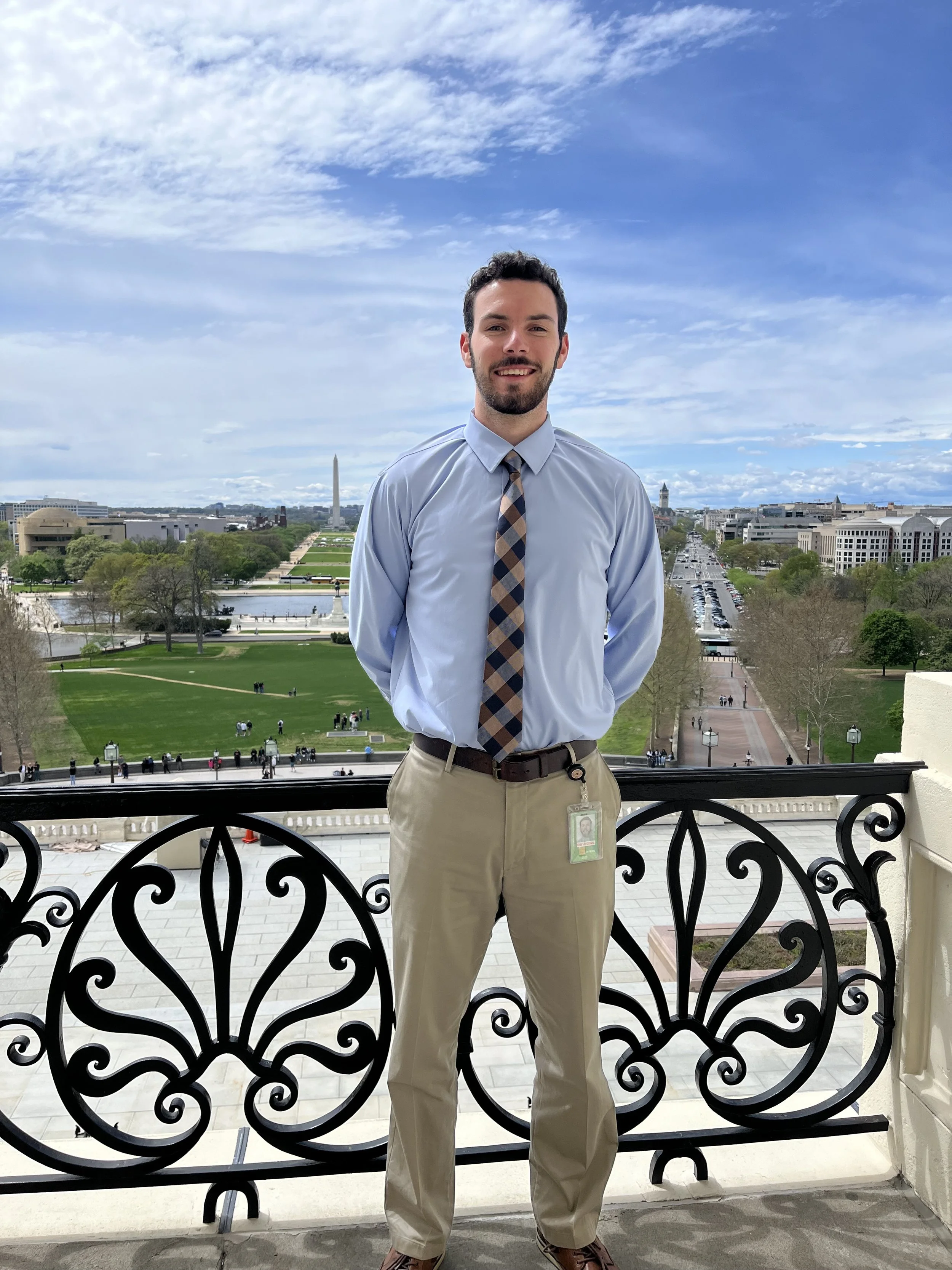 A man with dark hair and a beard, wearing a light blue dress shirt, beige pants, and a checkered tie, stands on a balcony with ornate black railing. In the background, the Washington Monument, the U.S. Capitol, and other city buildings are visible under a partly cloudy sky.