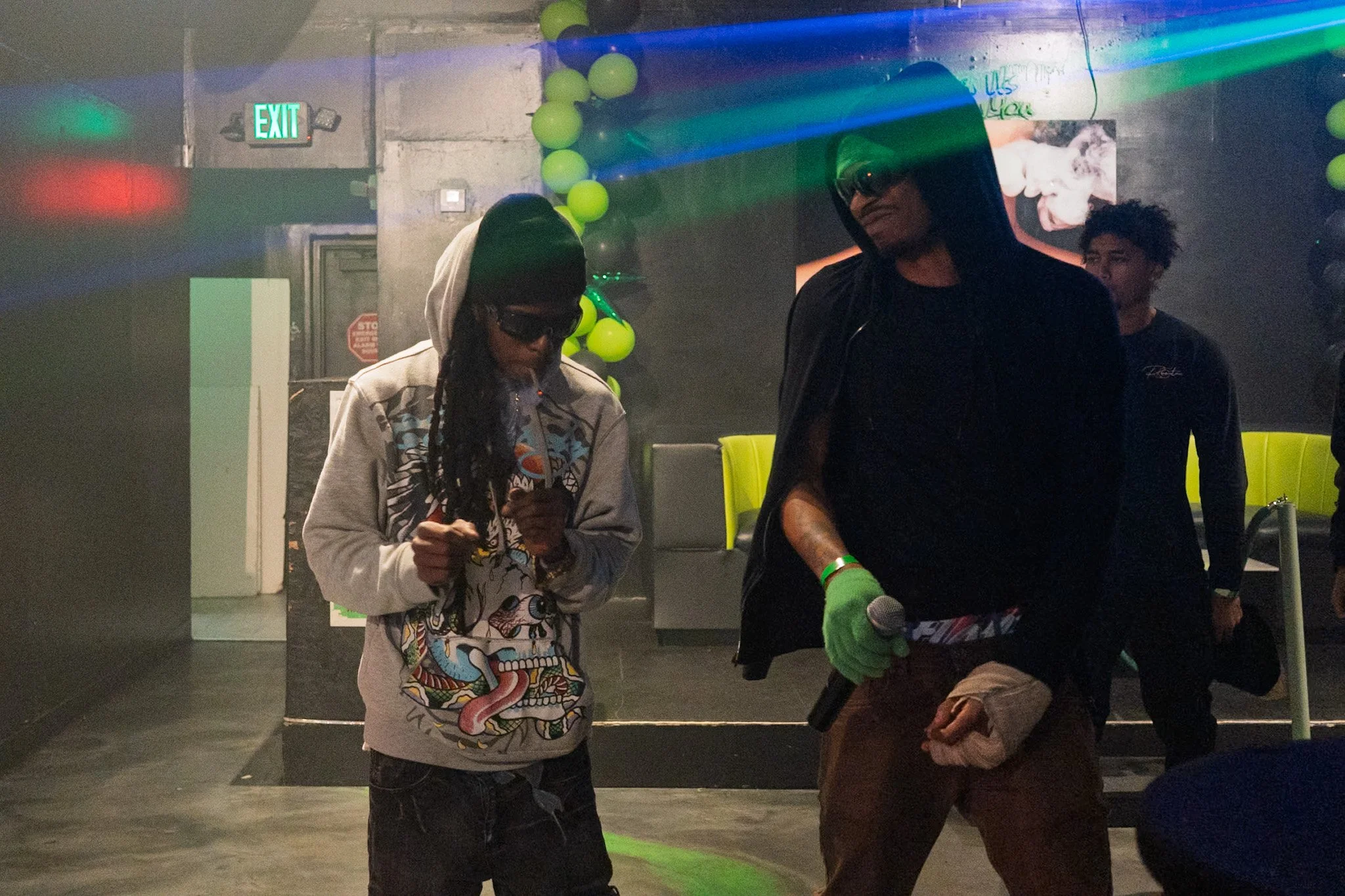 Two men are at a rave or party, with colorful lights and green balloons in the background. One man is wearing a hoodie with a skeletal design, and the other is wearing black sunglasses, a black hoodie, and gloves, holding a microphone.