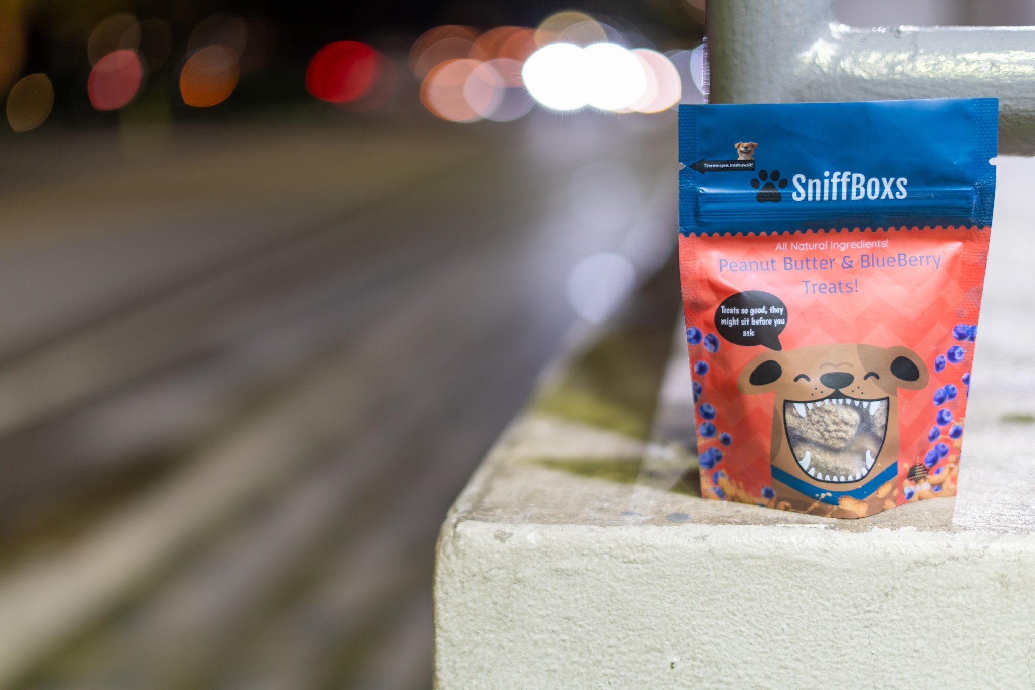 A bag of SniffBoxes treats for dogs, with a colorful bear graphic, placed on a concrete ledge near a wall, blurred lights in the background.