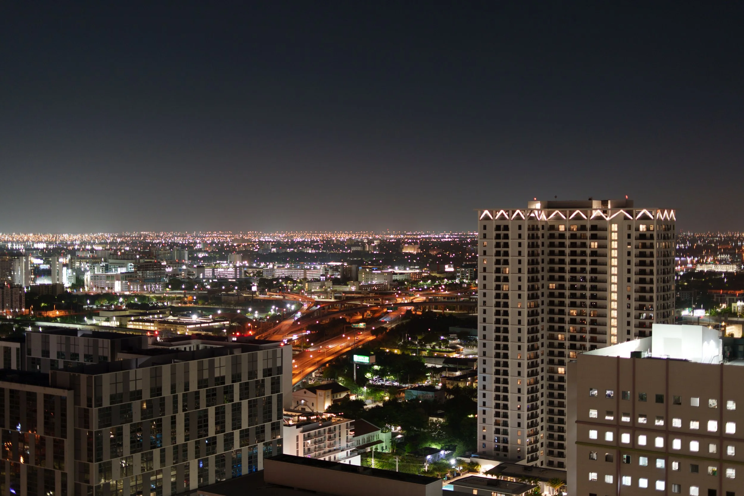 Nighttime cityscape view from a high-rise building showing illuminated skyscrapers and streets with lights and traffic, with a dark sky overhead.