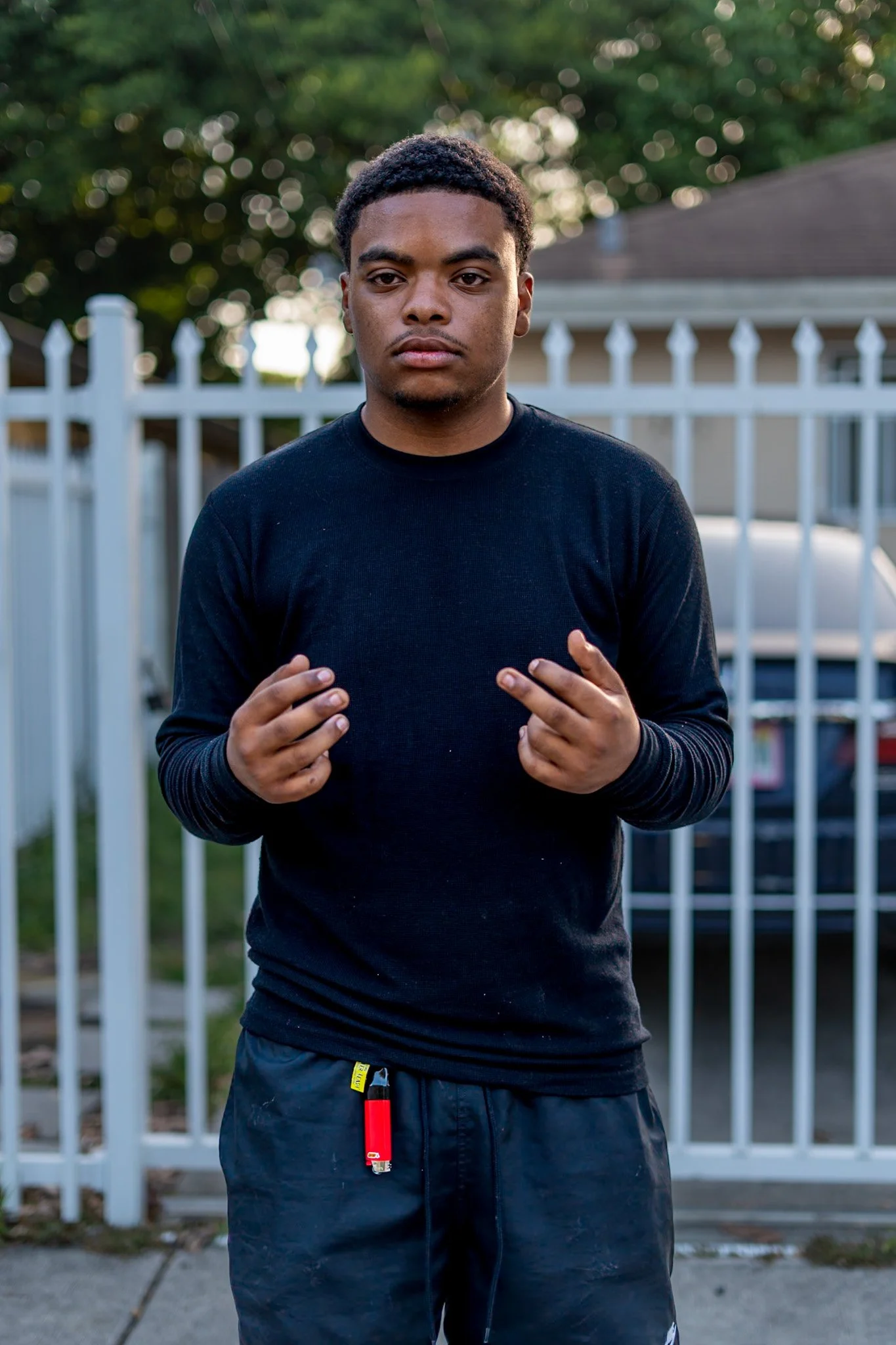 Young man with curly hair wearing a black long-sleeve shirt and dark shorts, standing outdoors in front of a white picket fence and green trees, looking directly at the camera.