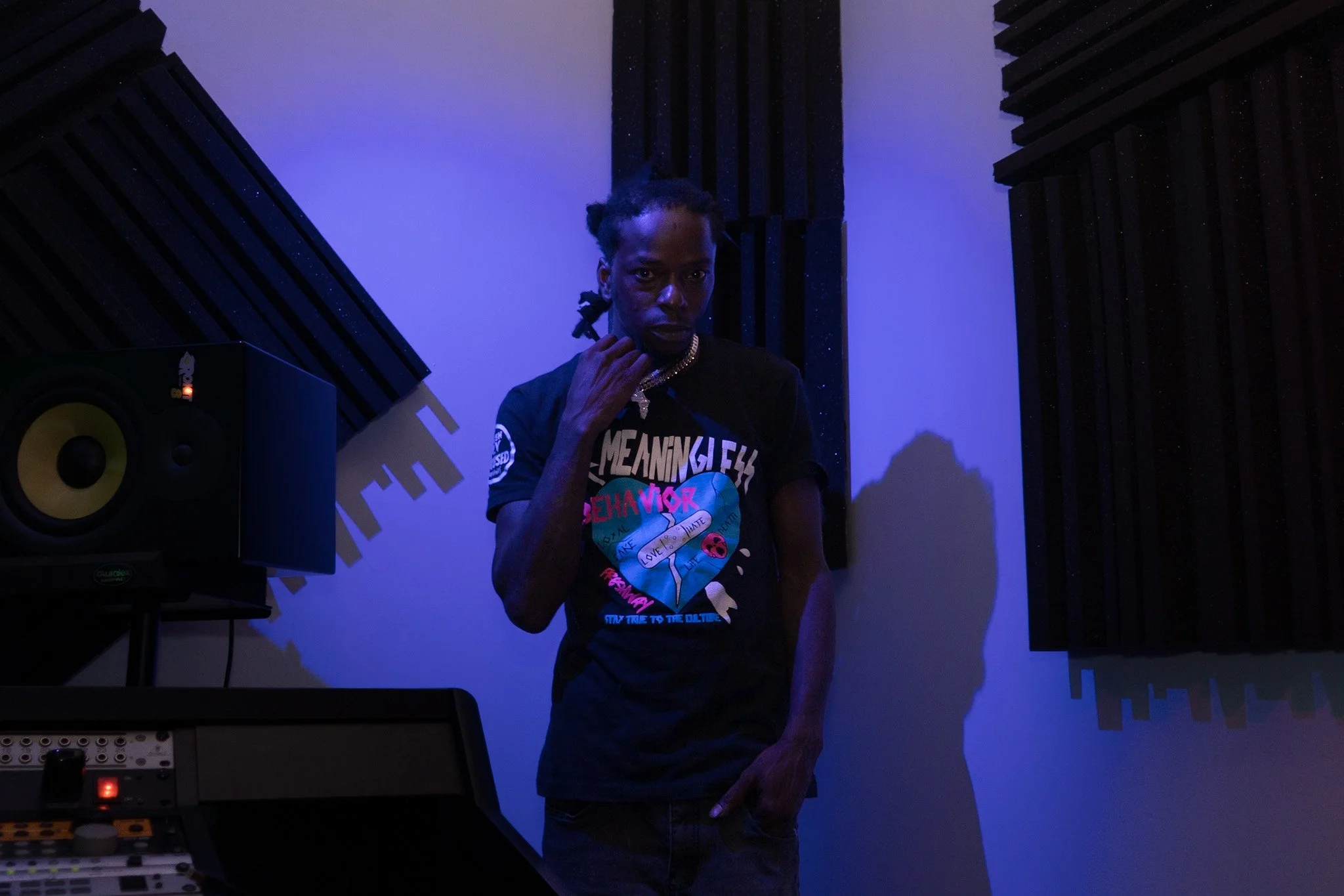 A person standing in a dark recording studio wearing a T-shirt with colorful text and graphics, with music equipment and black soundproofing foam on the walls.