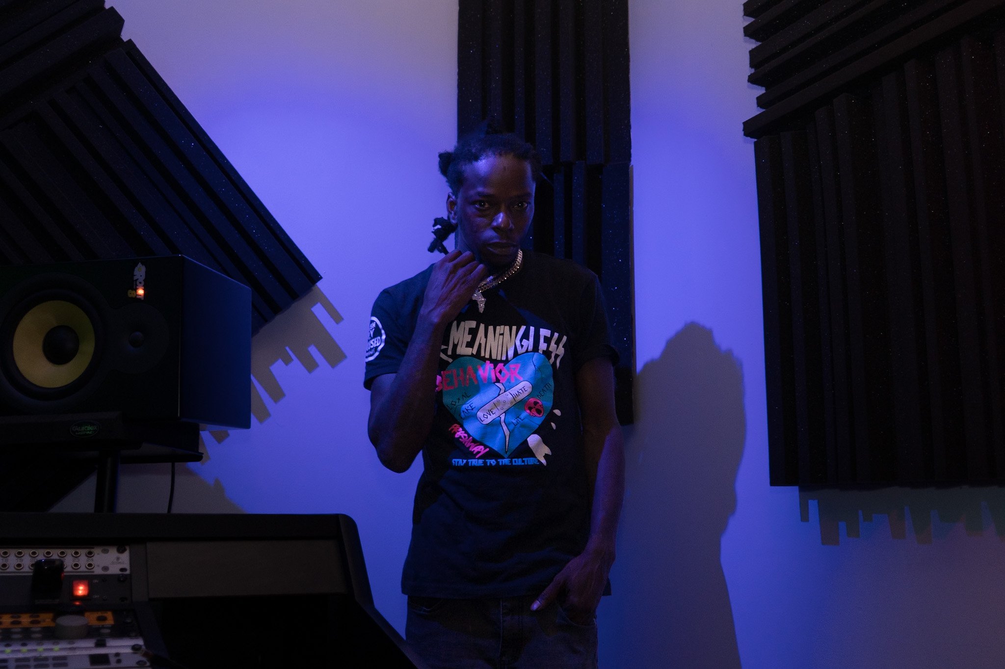A person with braided hair, wearing a black t-shirt with colorful graphics, wears a gold chain necklace, and stands in a dimly lit recording studio or music production room, surrounded by speakers and soundproofing panels.