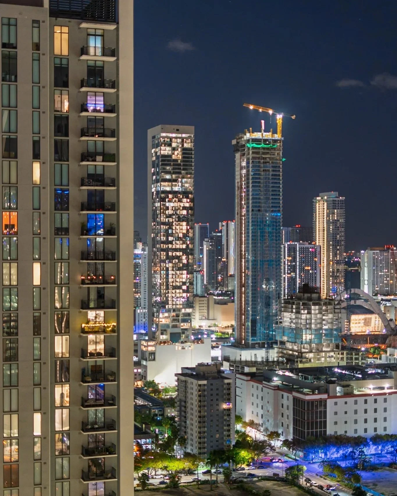 Nighttime cityscape with illuminated high-rise buildings under a clear dark sky, some buildings still under construction with cranes, and lined streets with parked cars and illuminated trees.