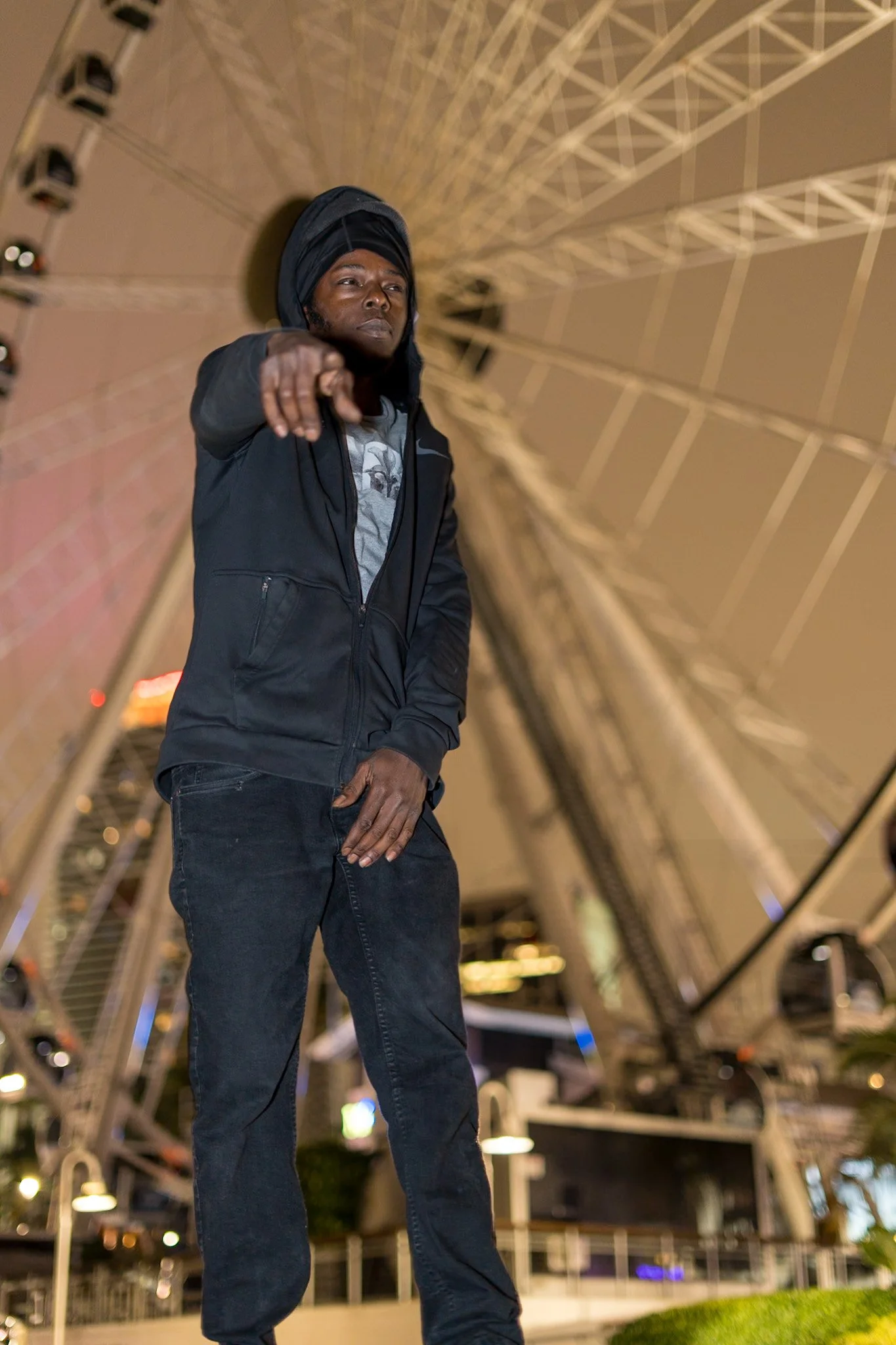 A young man is standing outdoors at night in front of a large Ferris wheel, pointing towards the camera. He is wearing a black hoodie, black pants, and a headscarf, with a serious expression.