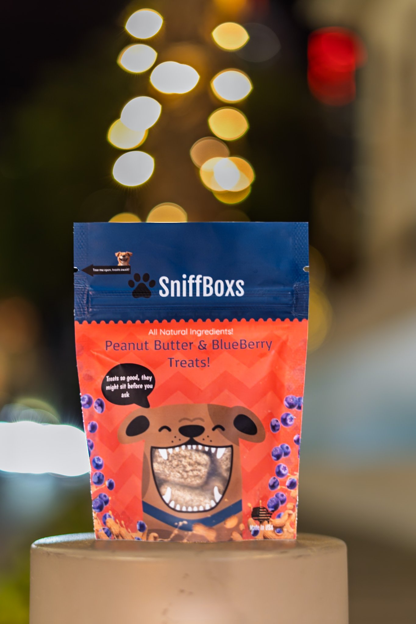 Close-up of a package of SniffBoxes peanut butter and blueberry dog treats with a cartoon dog face on the packaging, blurred bokeh lights in the background.