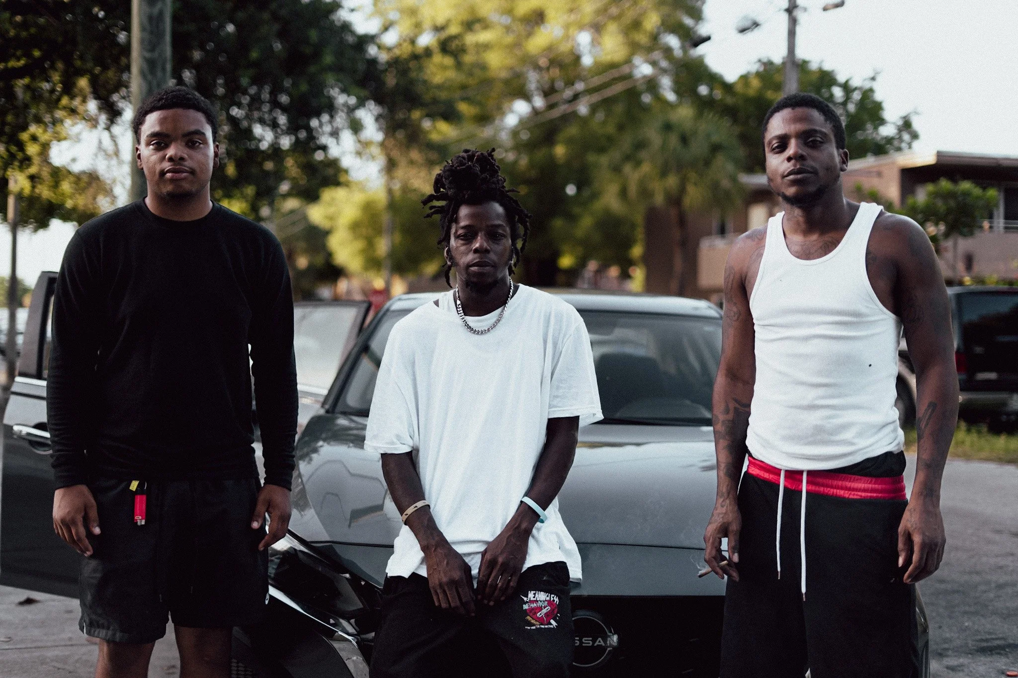 Three young men standing outdoors in front of a black car, with trees and a building in the background. They are dressed casually, with one wearing a black shirt, one in a white shirt, and one in a white tank top with red shorts.
