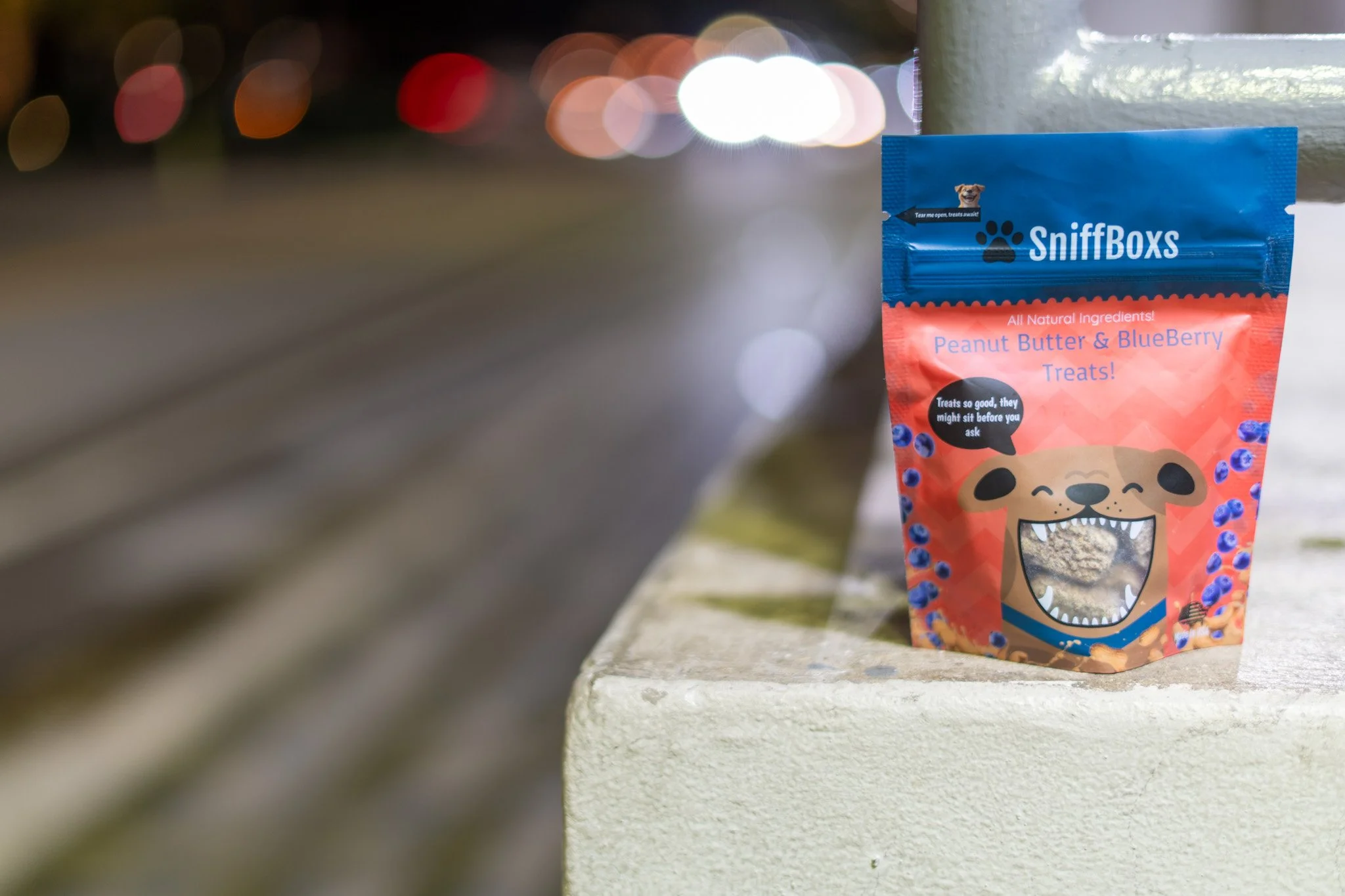 A bag of SniffBox dog treats featuring a smiling cartoon dog with an open mouth and sharp teeth. The bag is labeled 'Peanut Butter & Blueberry Treats' and has a clear window showing the treats inside. The background is blurred with colorful lights, suggesting an outdoor setting at night.