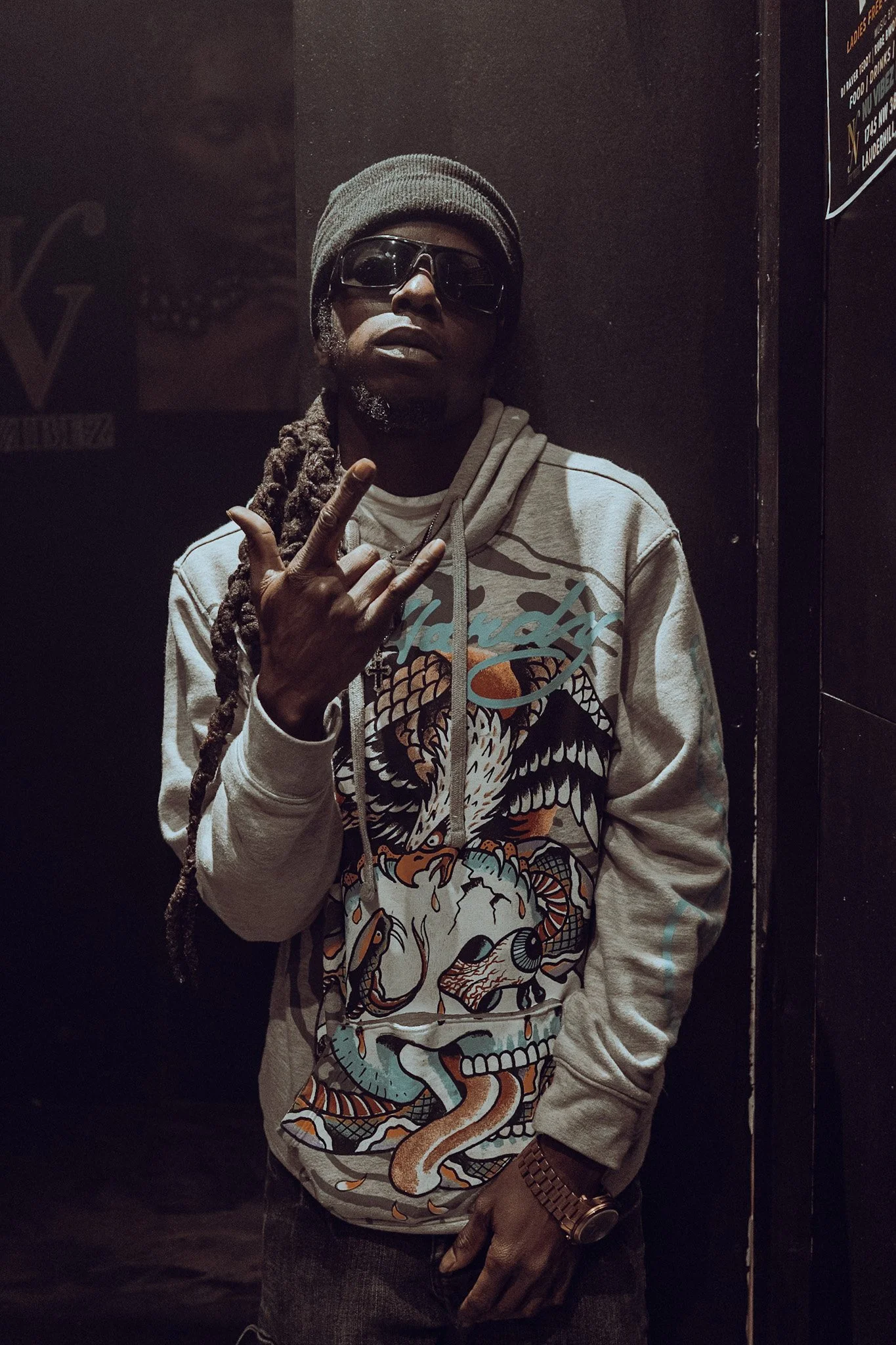 A man with long dreadlocks wearing a gray beanie, dark sunglasses, a light-colored hoodie with a graphic design of a dragon or similar creature, and a gold watch on his left wrist, standing against a dark wall and making a hand gesture.