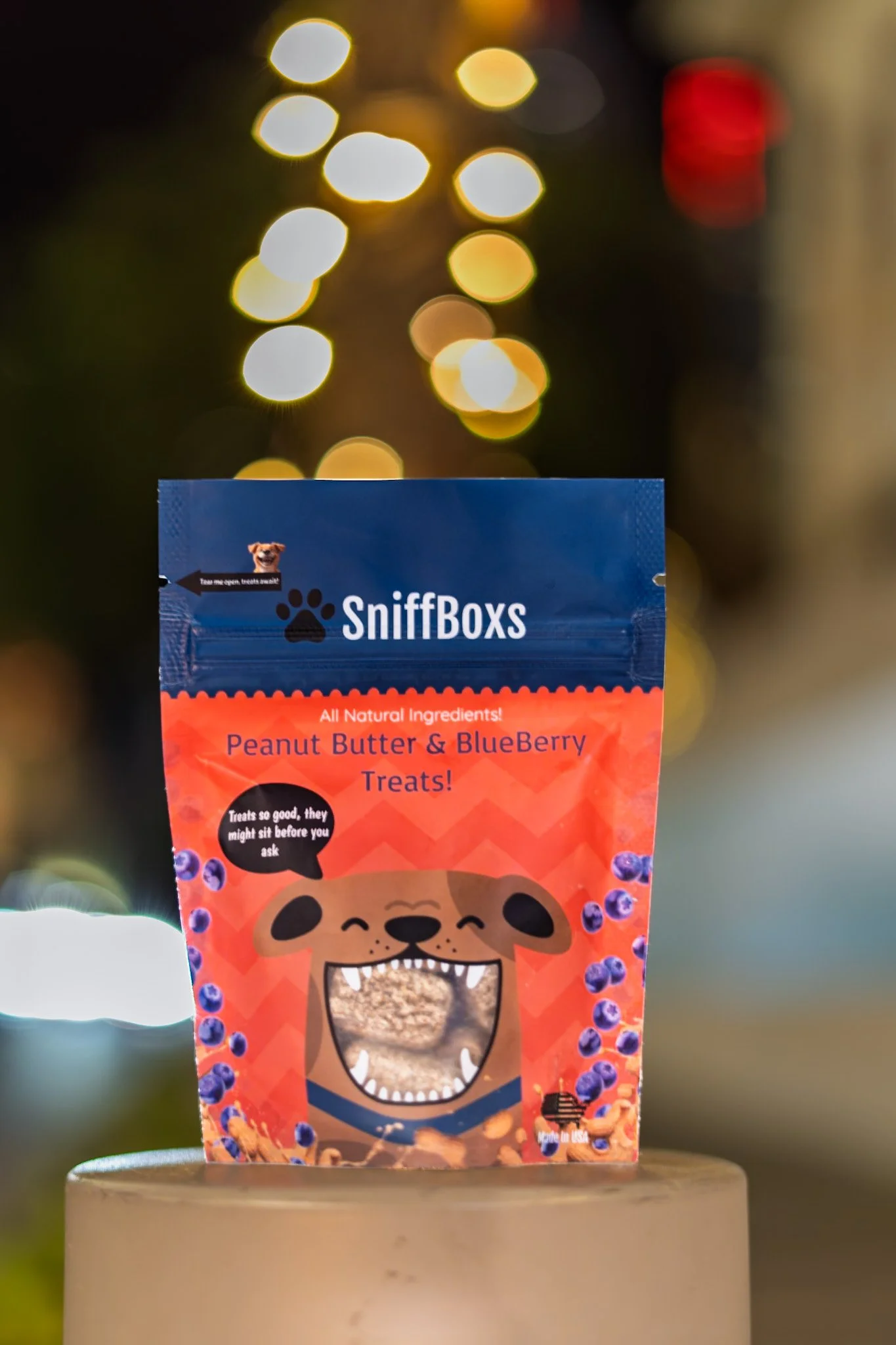 A bag labeled 'SniffBoxs' with a cartoon dog illustrating the treat's flavor, peanut butter and blueberry, with bokeh lights in the background.