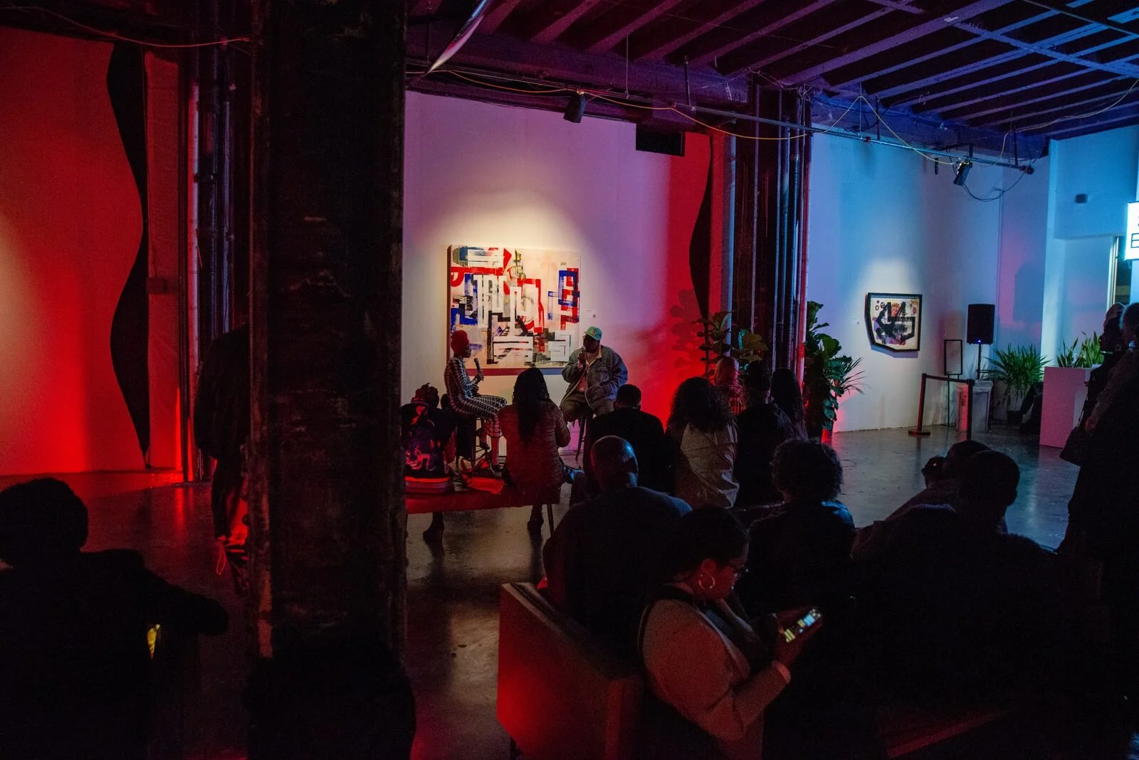 Scene from an art gallery or event space with an audience seated in front of a small stage where two people are having a conversation, one holding a microphone. The walls are decorated with abstract artwork, and the room features colorful lighting and potted plants.