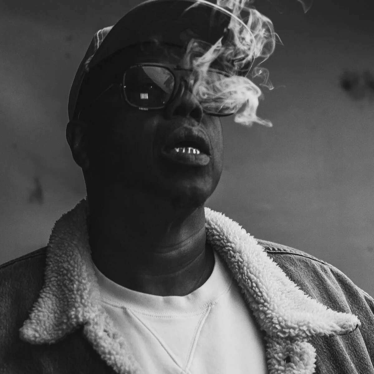 A black and white photo of a man wearing glasses and a cap, smoking with smoke wafting around his face. He is dressed in a denim jacket with a shearling collar and a shirt underneath.