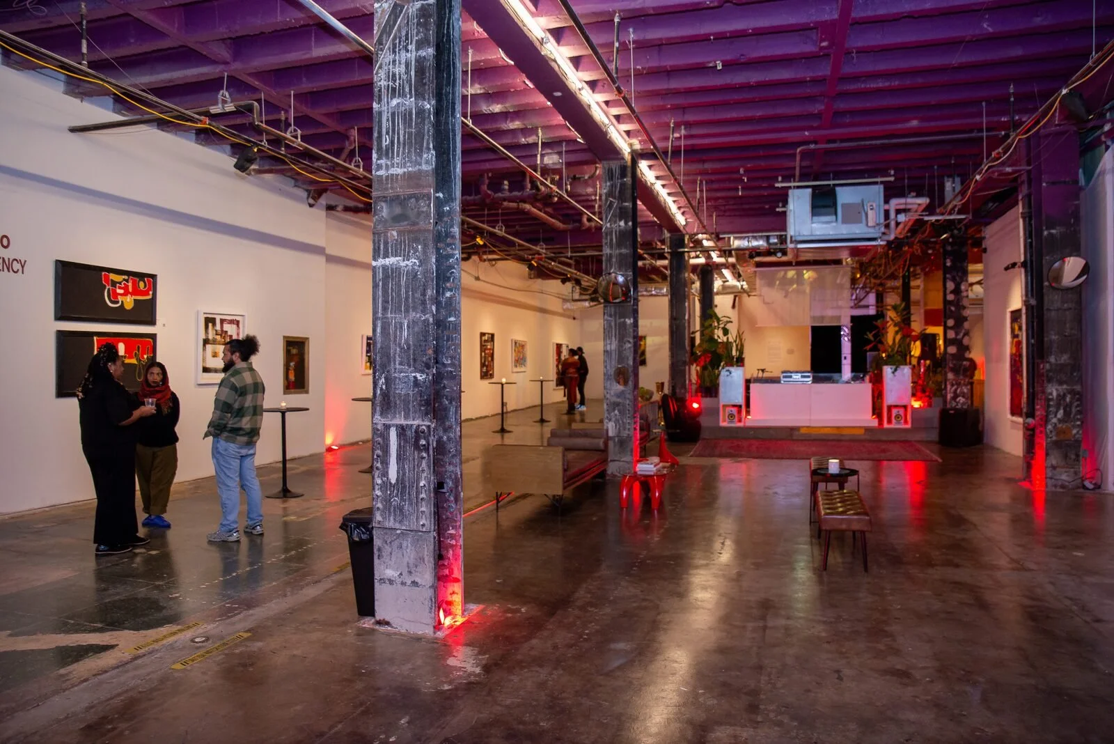 An art gallery with visitors, illuminated by red and purple lighting, featuring various artworks on white walls, and a DJ booth at the back with speakers and plants, in an industrial space with exposed pipes and concrete floors.