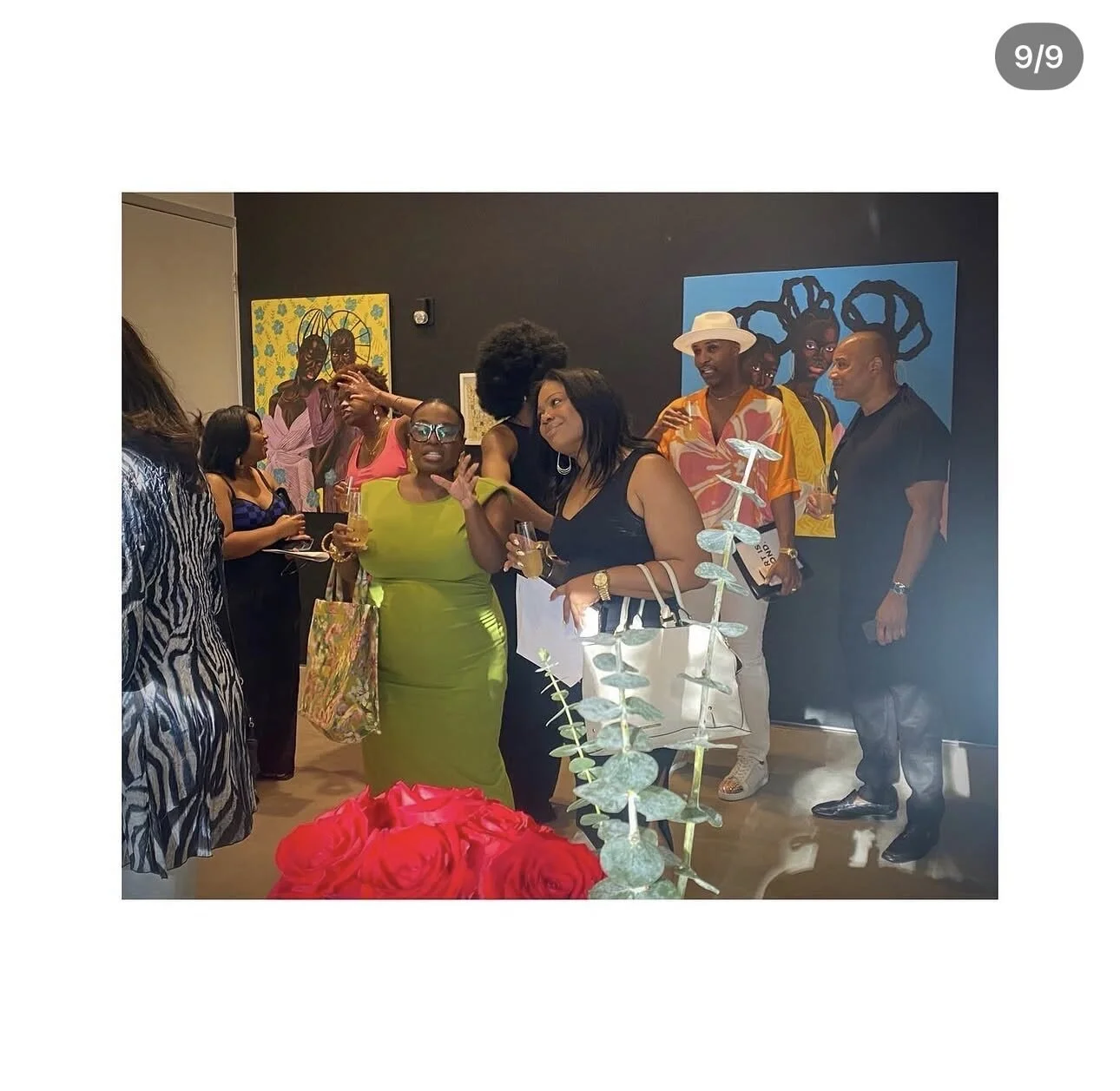 Group of women and men at an art gallery opening, engaging in conversations, with colorful artwork on the black wall behind them.