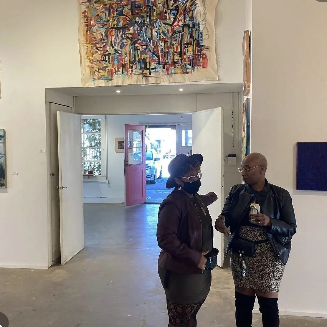 Two women having a conversation inside an art gallery, with artworks hanging on the walls and a view of the street outside through the door.