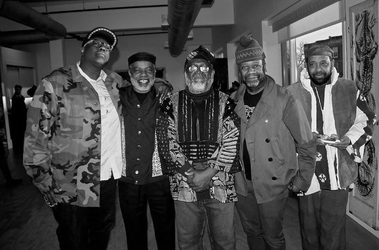 Group of five men standing together indoors, smiling, with artwork on the wall behind them, in a casual social setting.