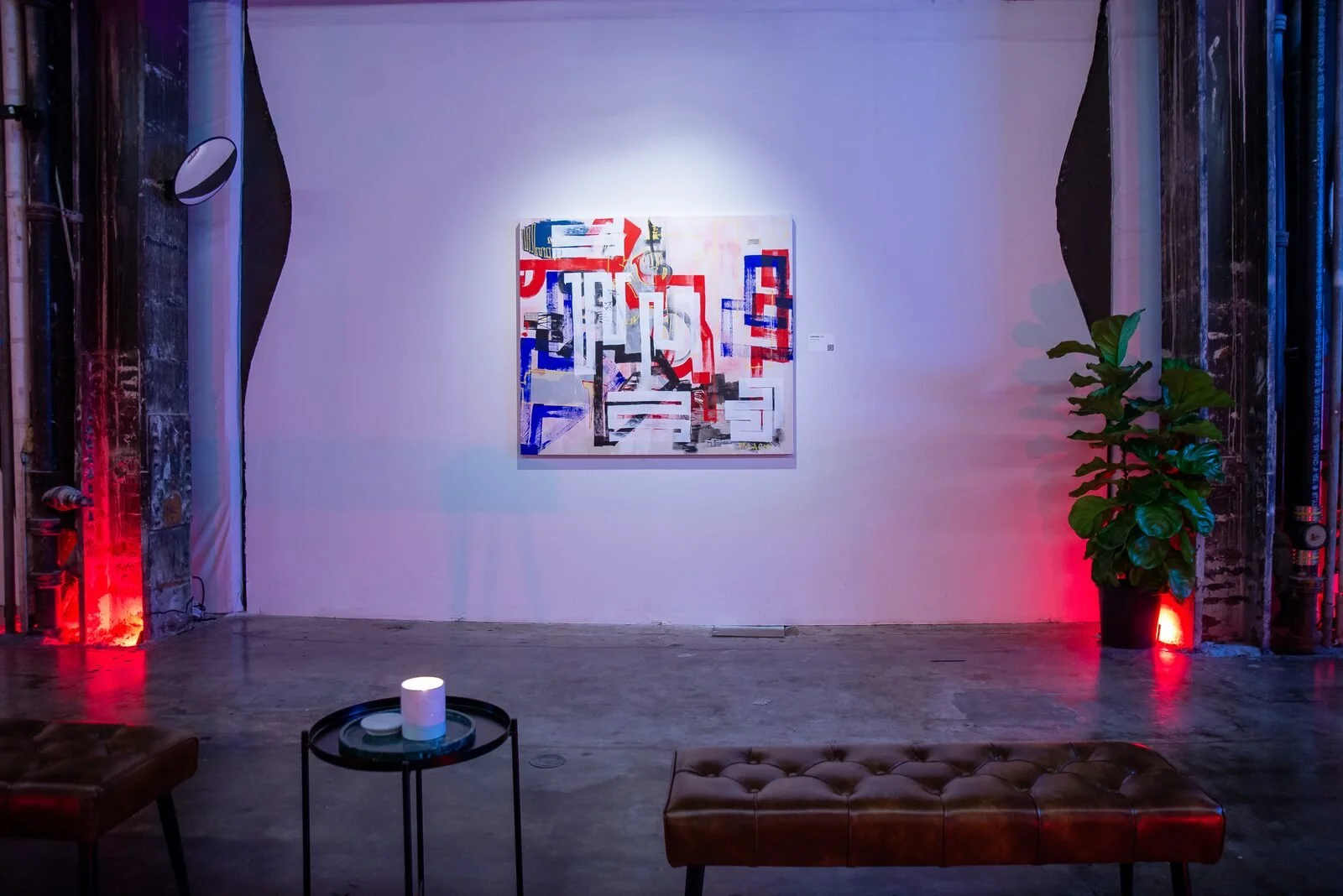 Art gallery with an abstract painting on the white wall, decorative lighting on brick columns, a green potted plant, a small table with a candle, and seating furniture in a cozy exhibition space.