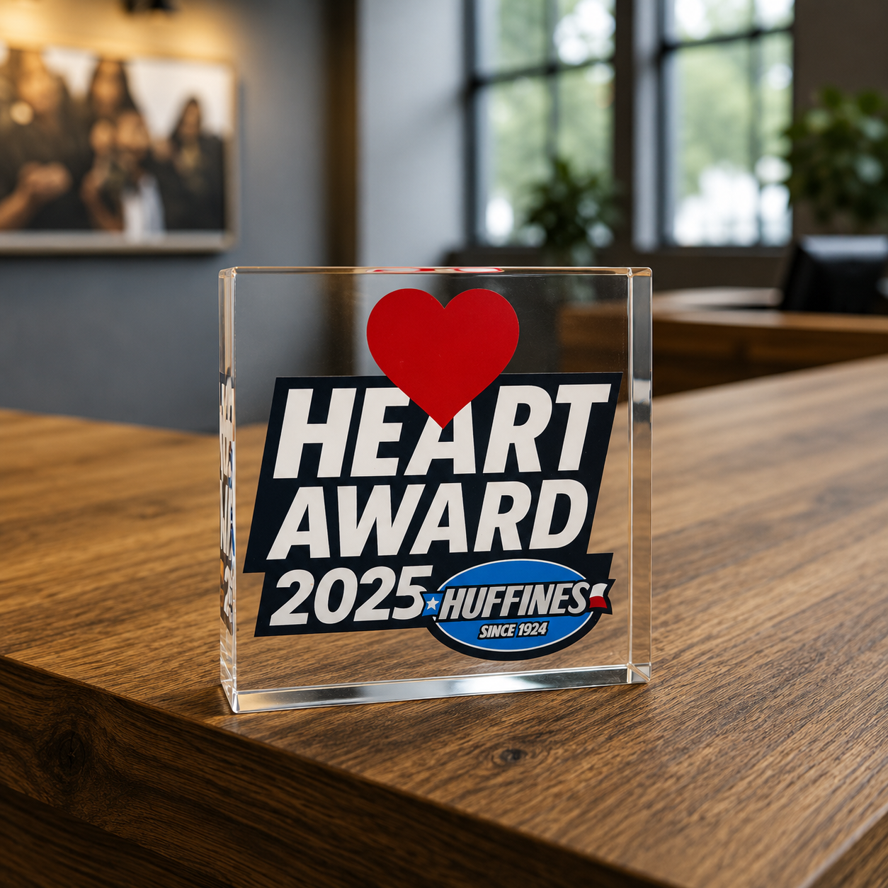 A clear acrylic award with a red heart at the top, displaying the text "HEART AWARD 2025" and a logo for Huffines with the text "Since 1924," placed on a wooden table in a room with large windows and some blurred pictures and plants in the background.