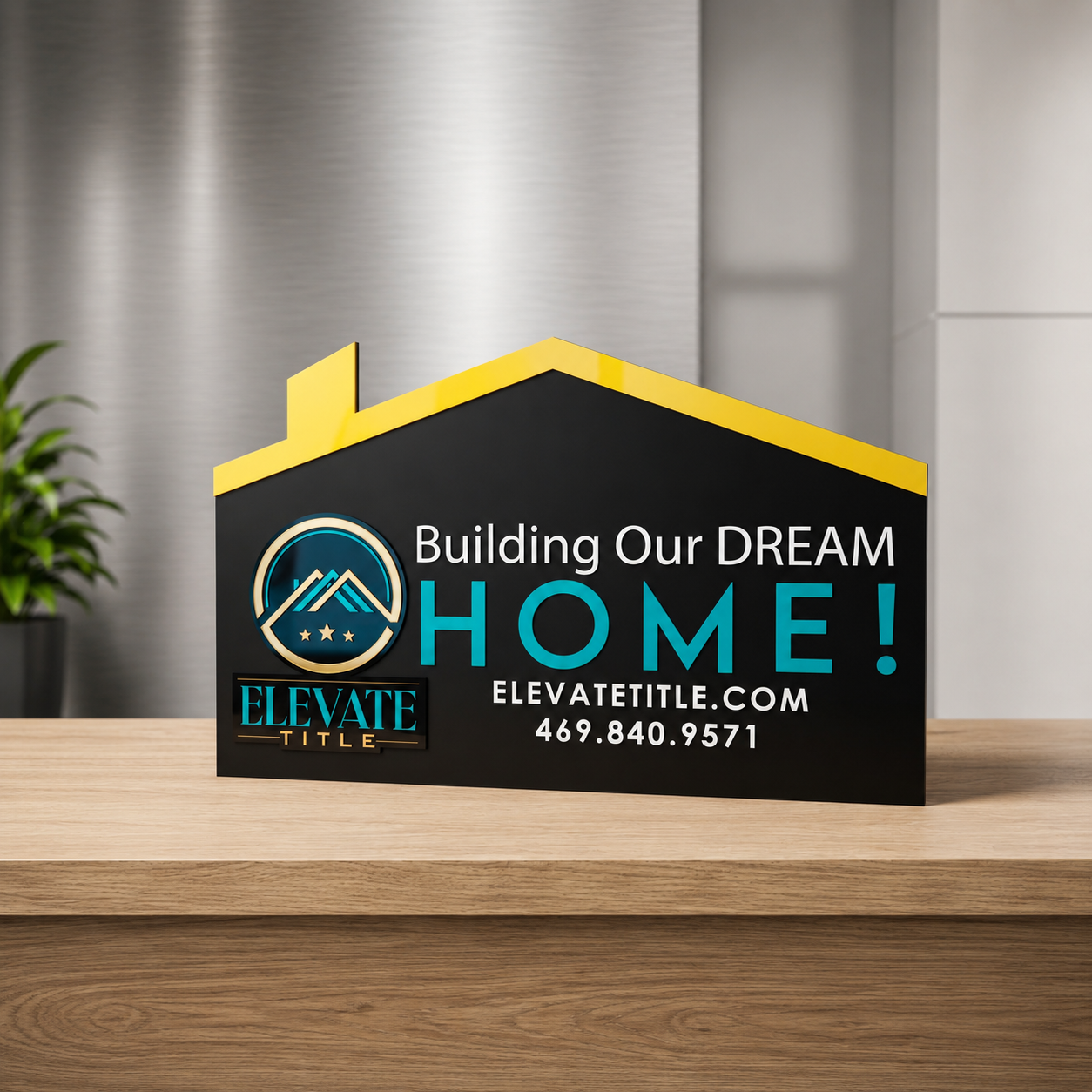 A promotional sign for a real estate company with the message "Building Our DREAM HOME!" and the company name "Elevate Title" along with contact details, placed on a wooden surface in an office setting.