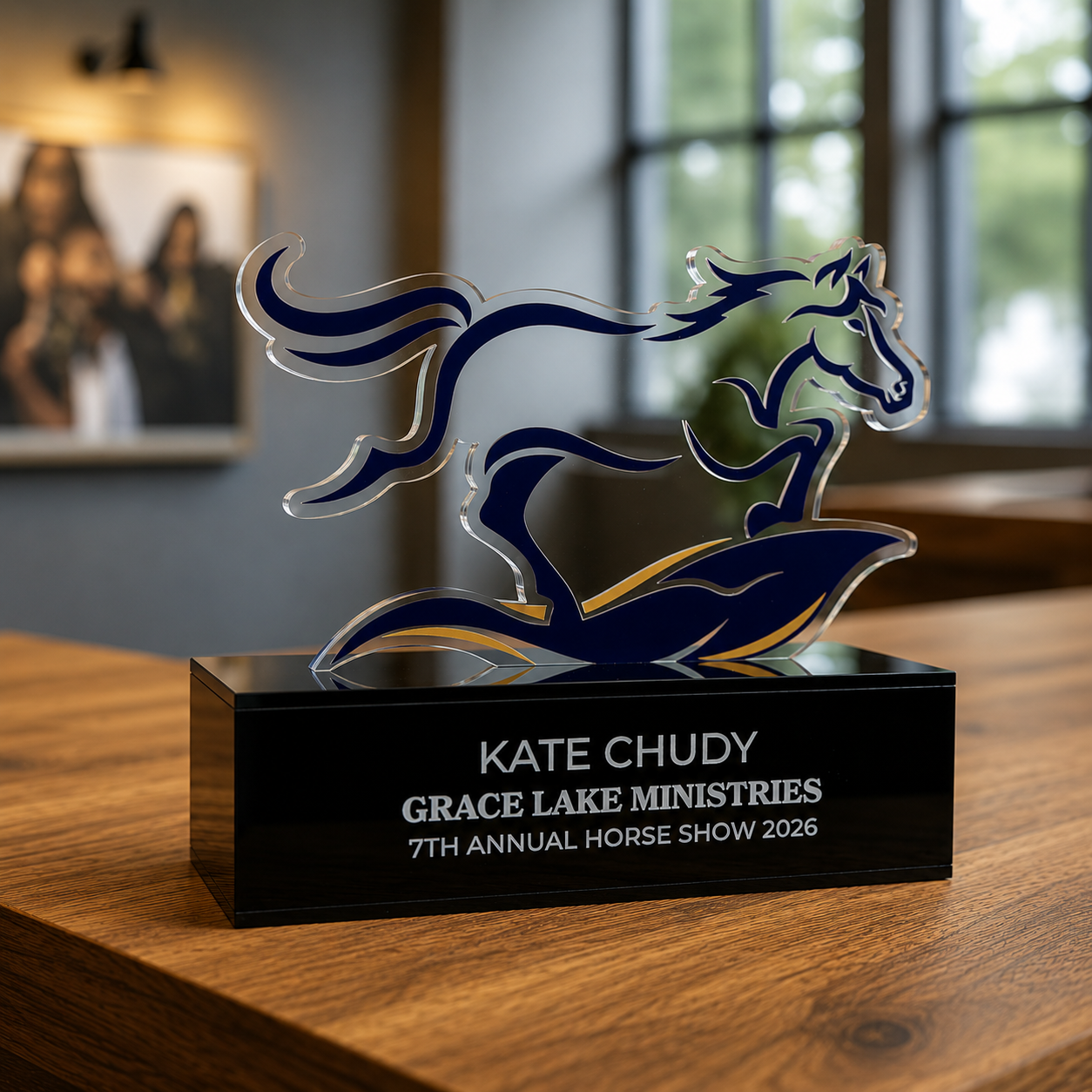 A glass horse trophy with blue accents on a black base, inscribed with the text 'Kate Chudy, Grace Lake Ministries, 7th Annual Horse Show 2026', sitting on a wooden table in a room with a window and blurred background.