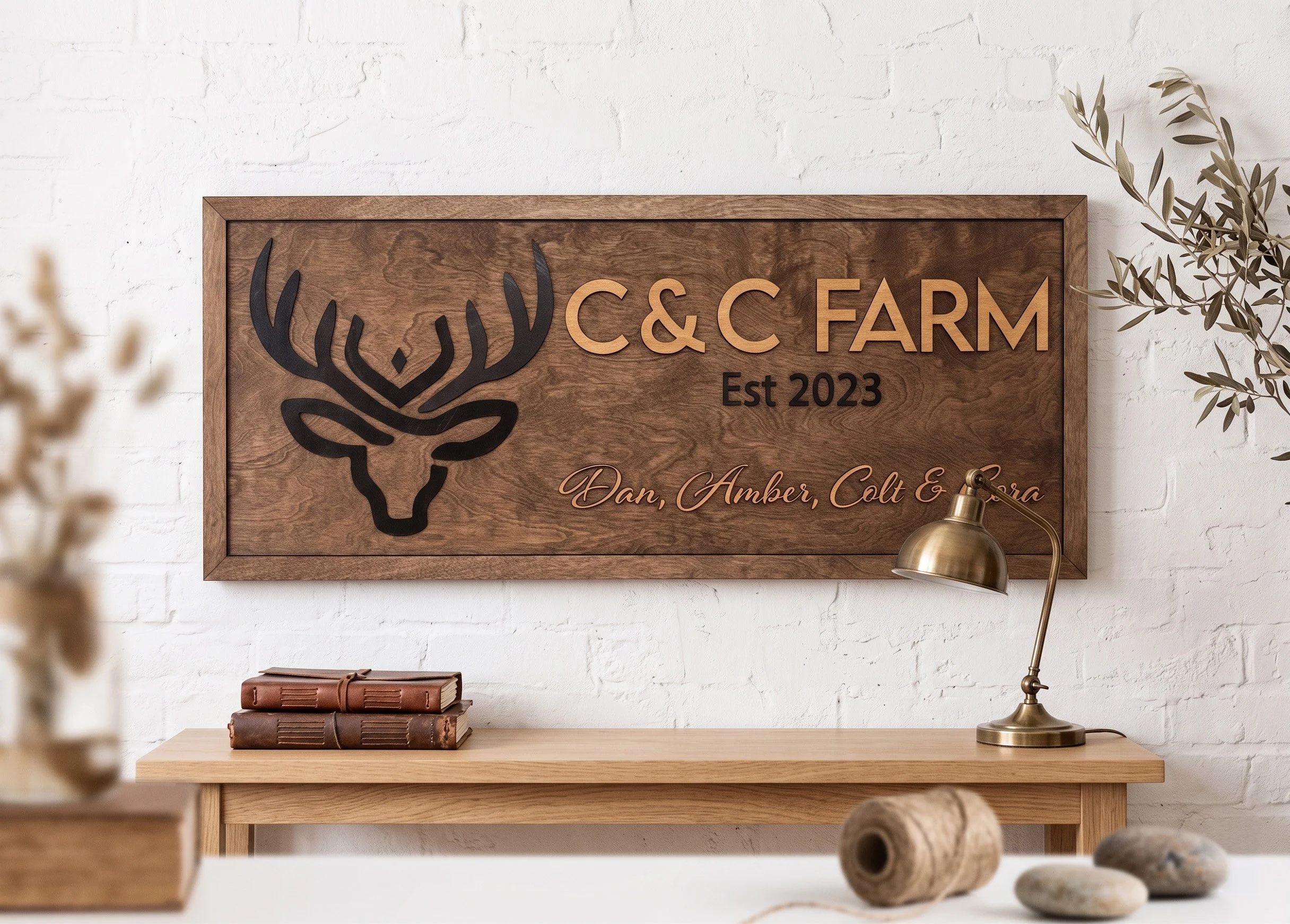 Wooden sign on white brick wall reads 'C&C FARM Est 2023' with a deer head silhouette. A brass desk lamp is on the right, and a stack of books is on the left, with some yarn balls in the foreground.