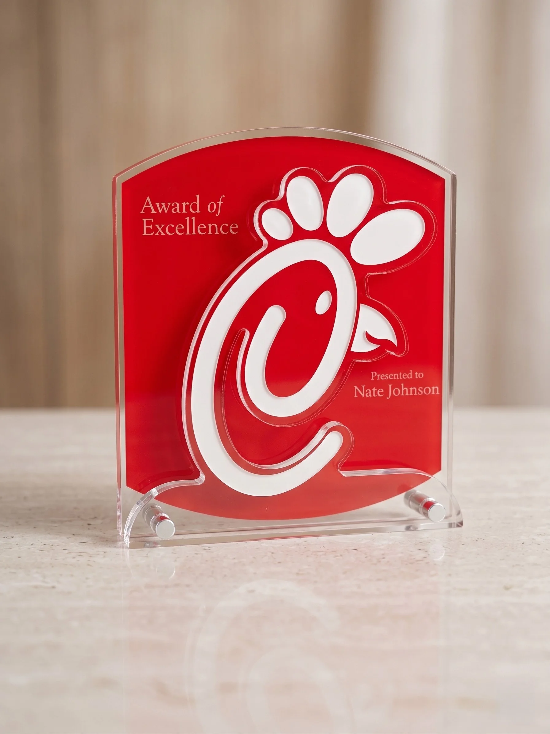 A red acrylic award plaque with the words "Award of Excellence" and "Presented to Nate Johnson" on it, placed on a clear stand on a beige countertop.