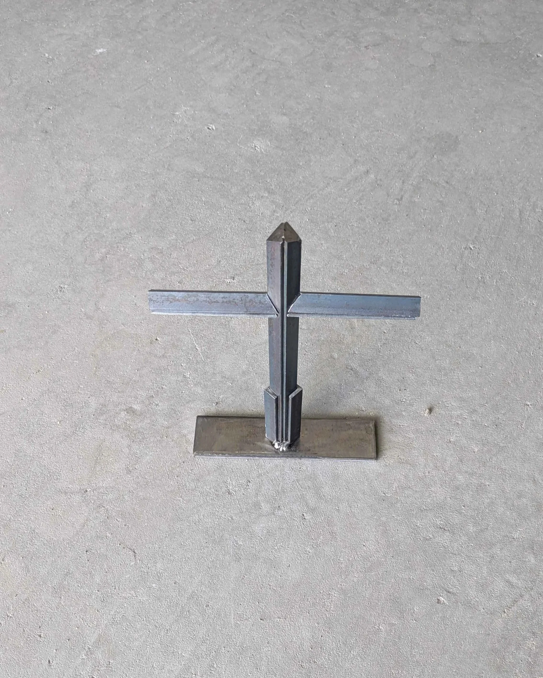 An overhead view of a metal art cross made from small pieces of angle iron placed on a stand, laying upon a slab of concrete..