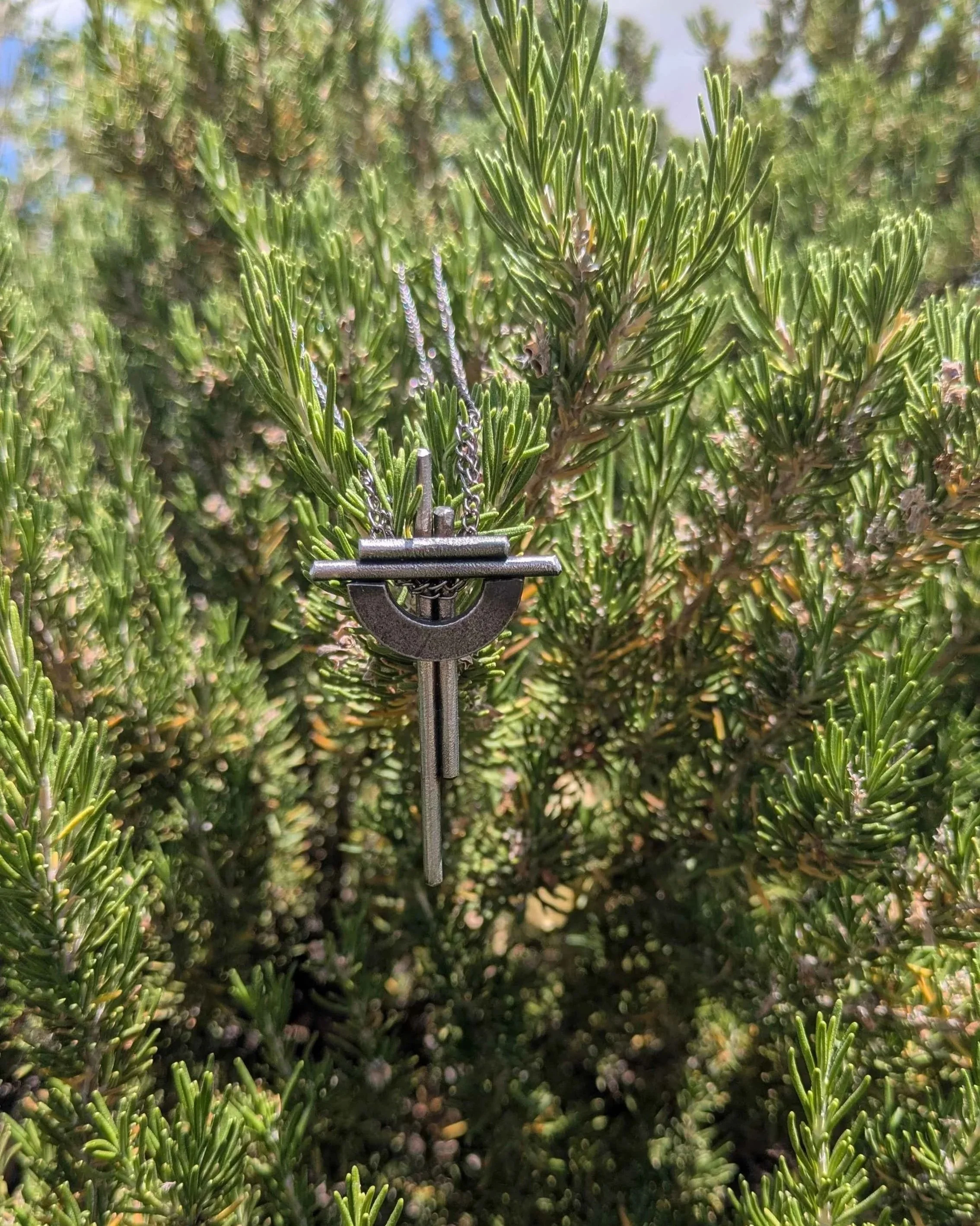 A handmade metal cross necklace with a chain, hung on a plant.