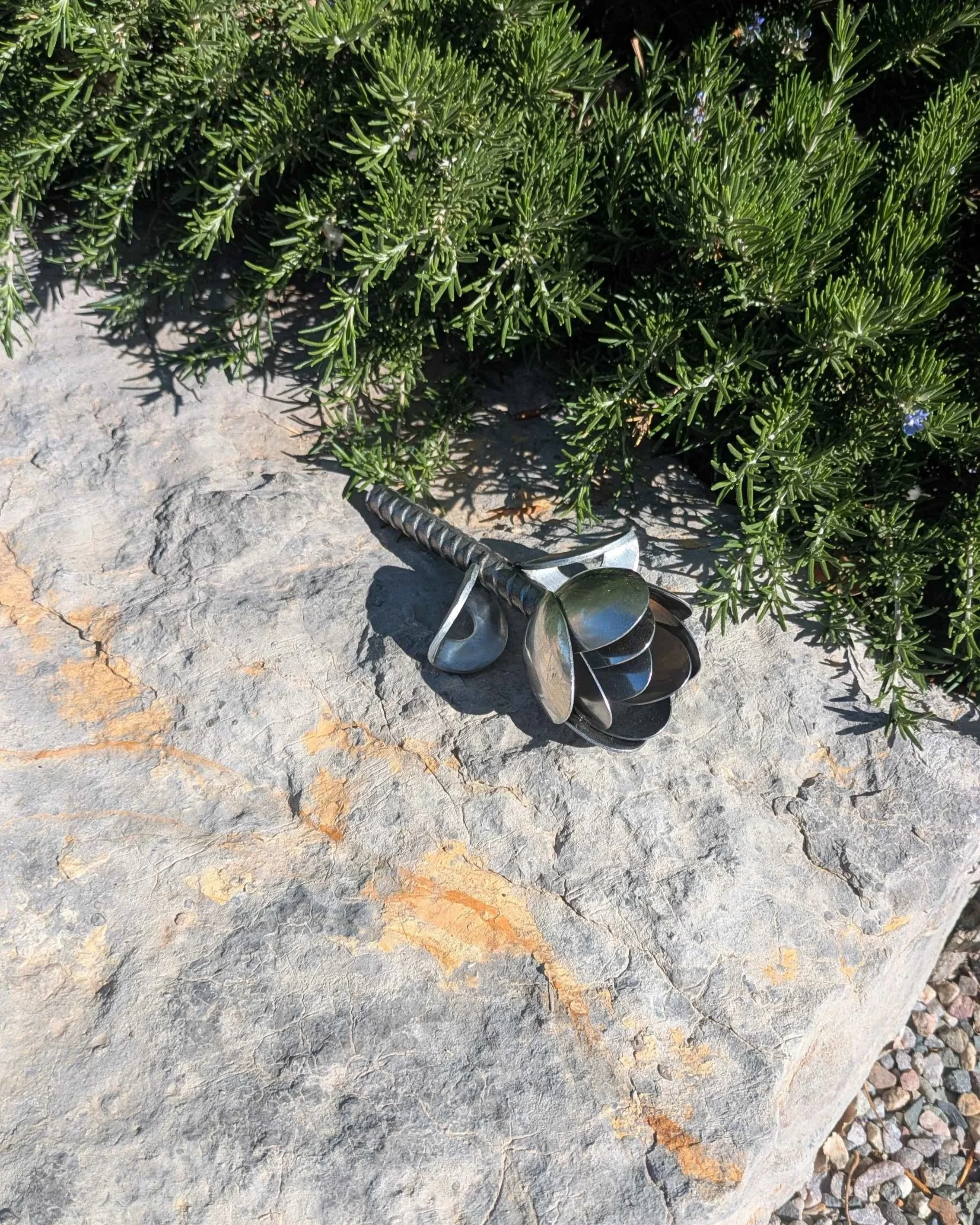A metal art tulip made from spoons, rebar and washers, sitting on a rock next to a bush.