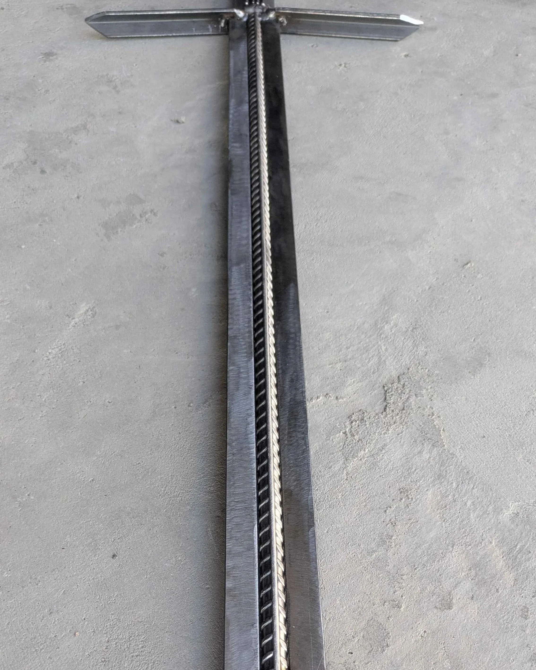 The blade of a metal art sword made from industrial style materials, laying upon concrete for a close-up picture.
