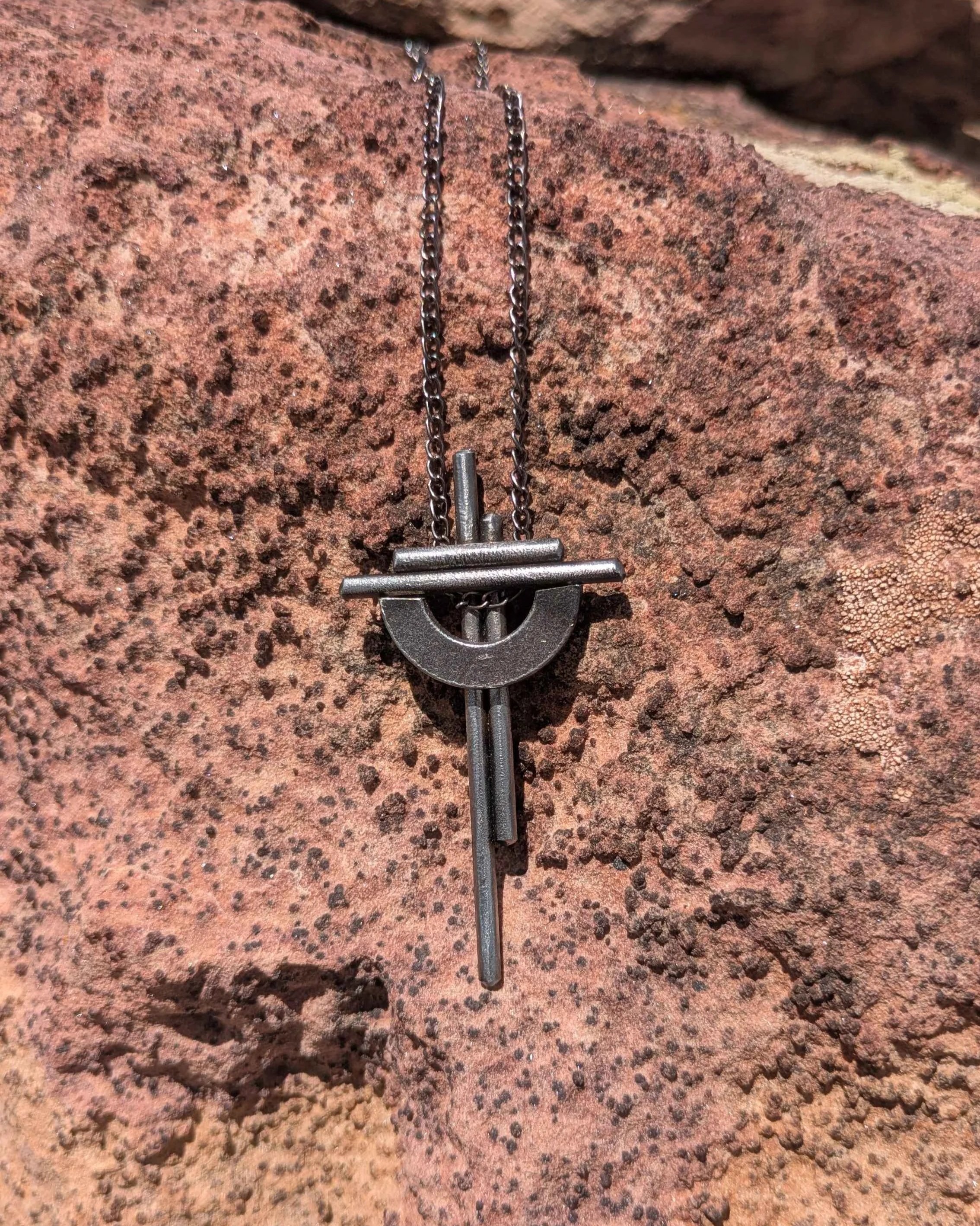 A handmade metal cross necklace with a chain, hung on a rock.