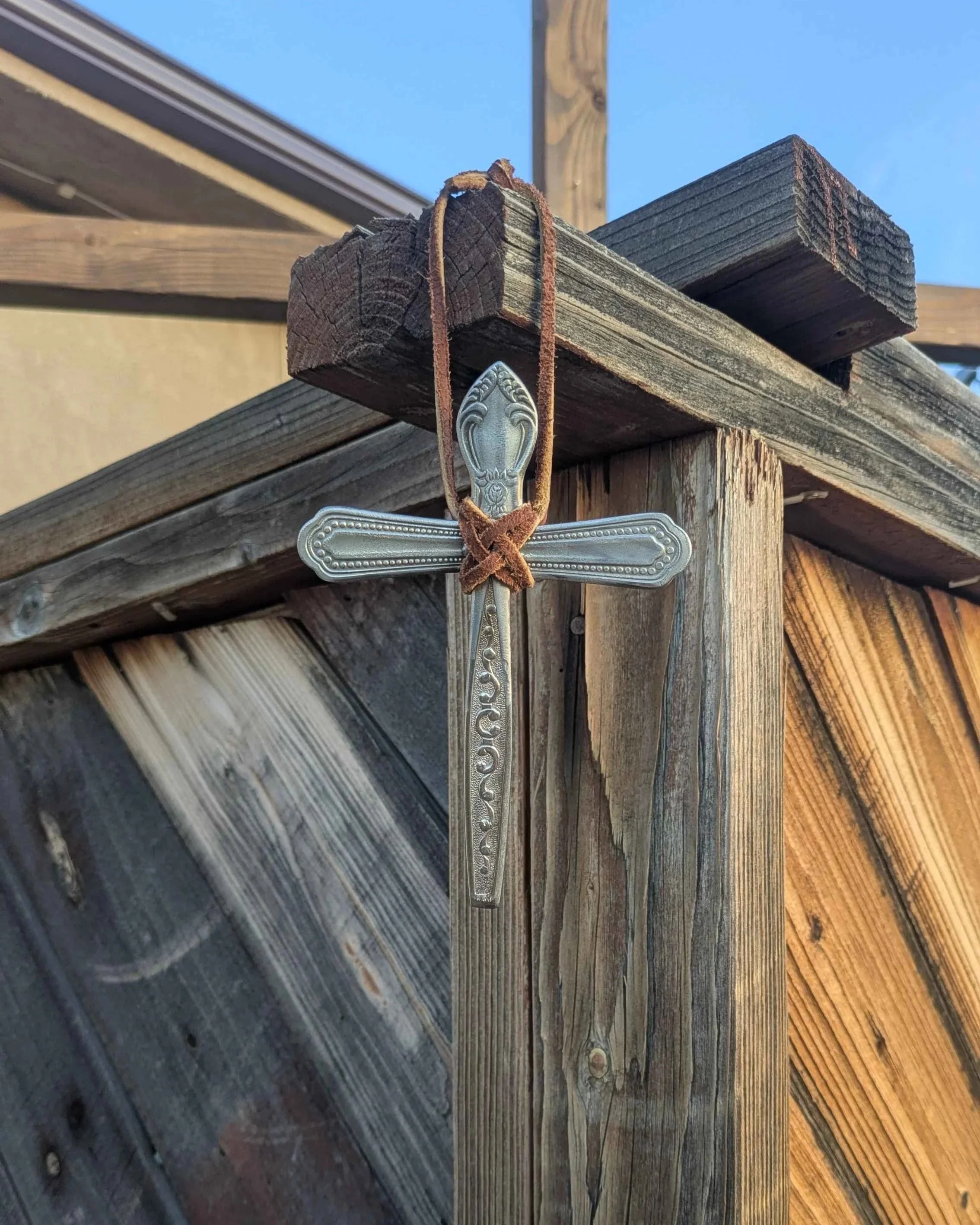 A silverware cross with an intricate leather hanging strap hung from a fence.