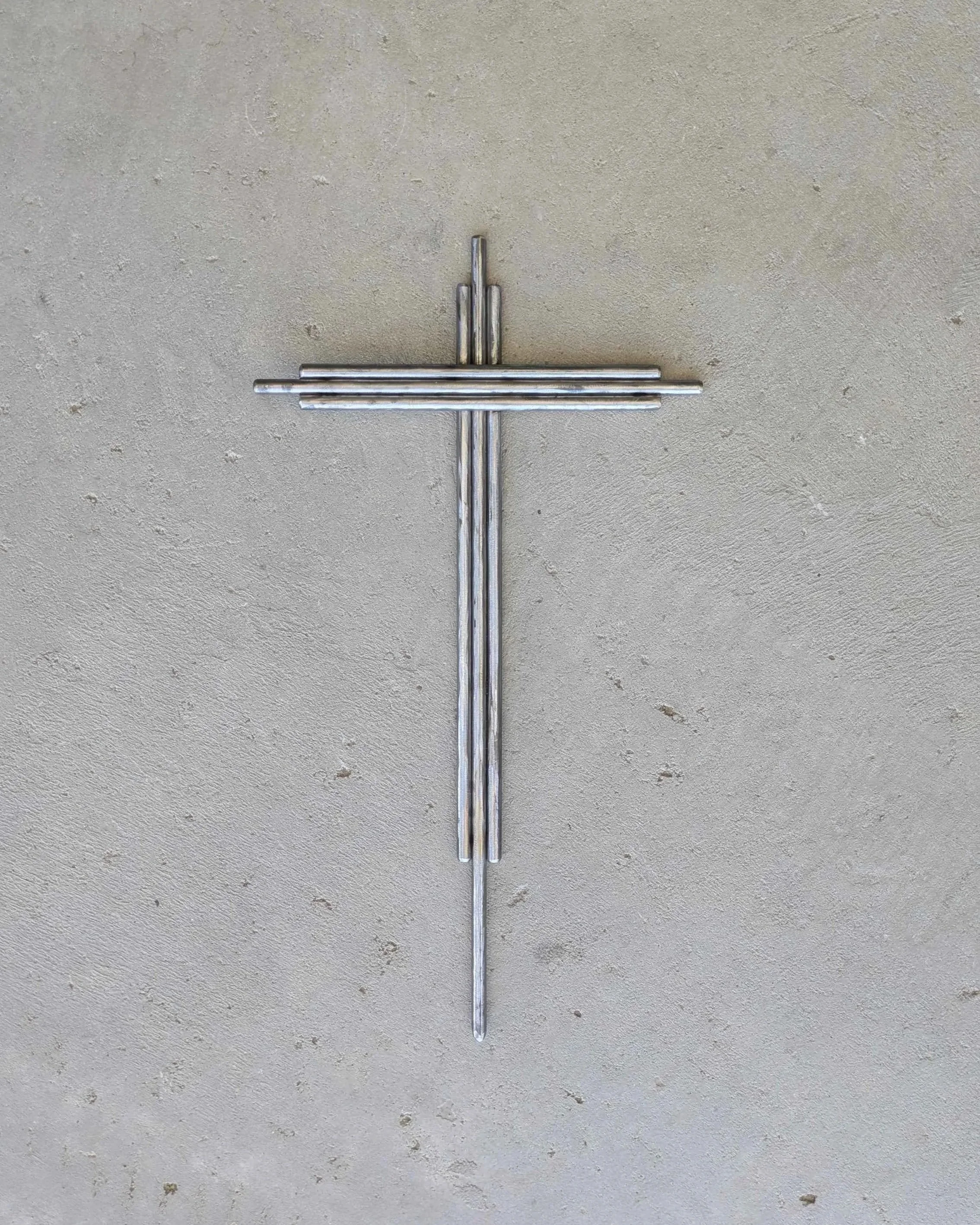 A metal art cross made from pieces of thick polished metal rod, laying on a slab of concrete.