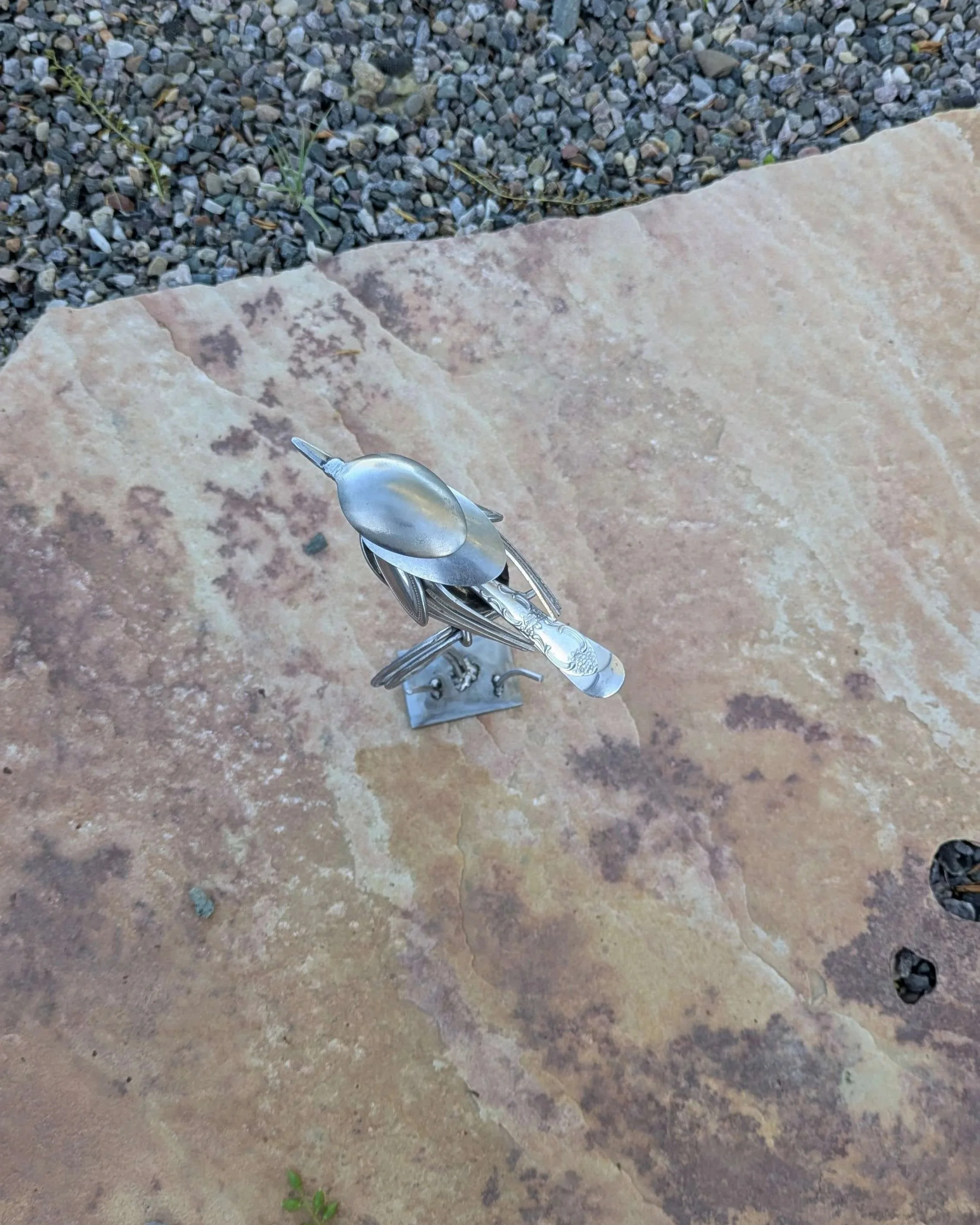 A handmade metal art bird sculpture made from recycled silverware sitting on a wooden shelf, resulting in a great piece of home decor.