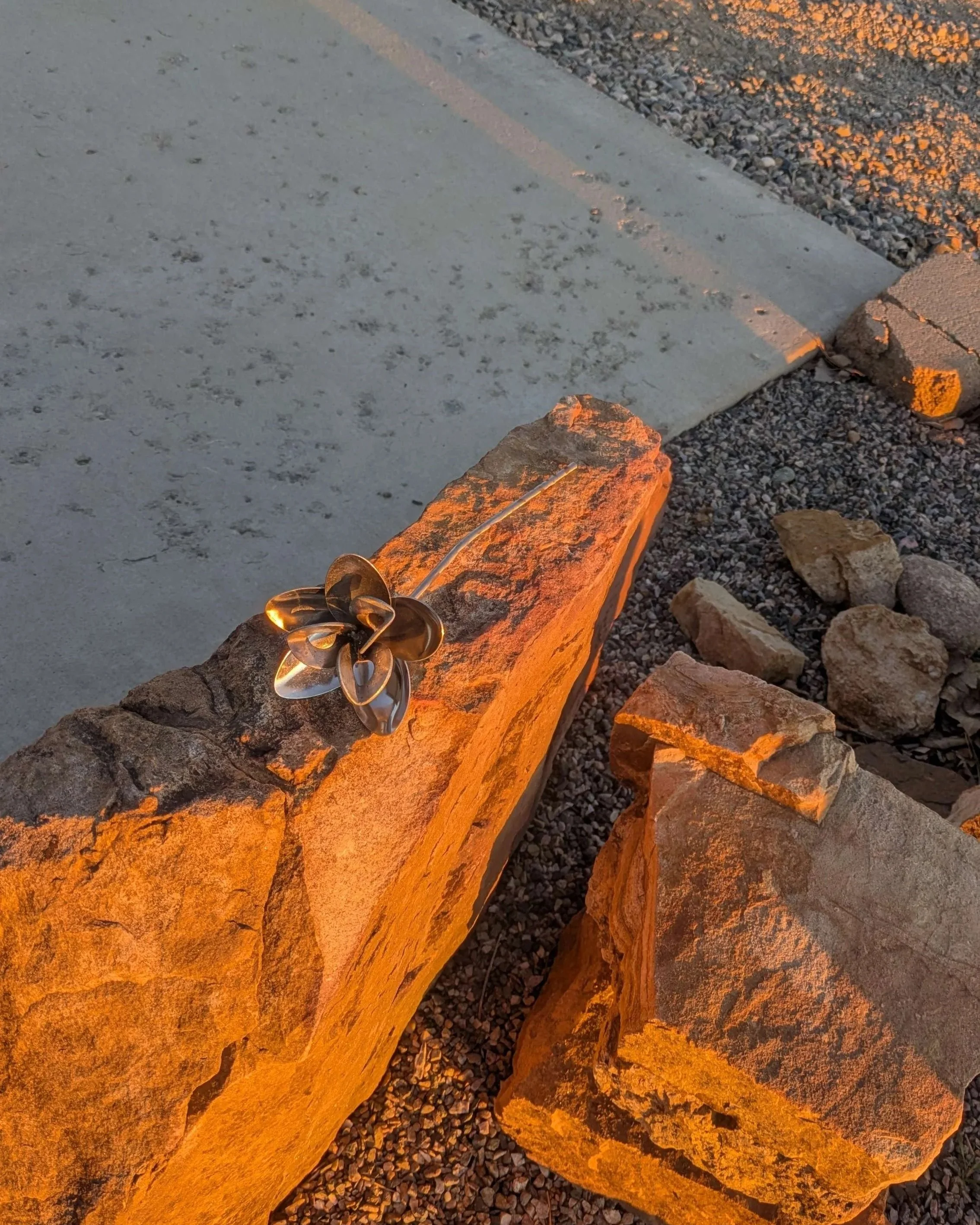 A metal art flower made from bent spoon heads, washers, and metal rod sitting on a rock.