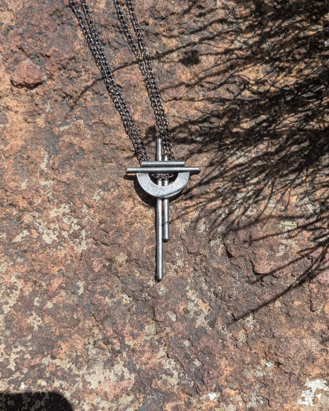 A handmade metal cross necklace with a chain, laying on a rock.