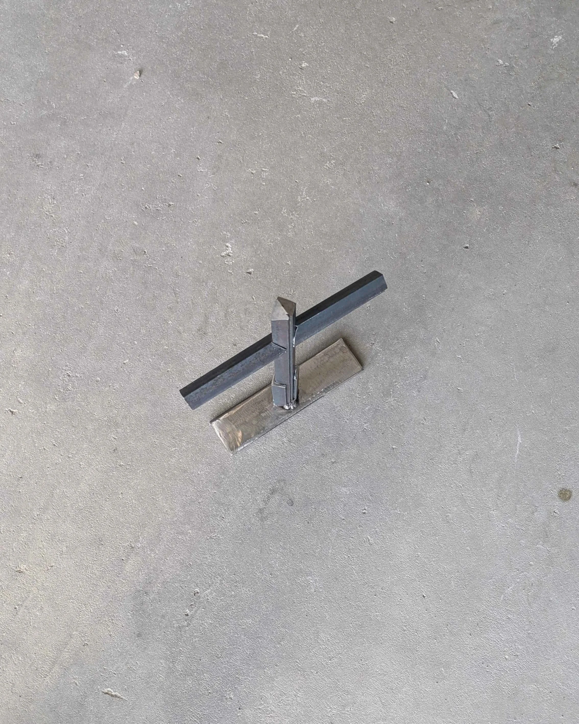 An isometric view of a metal art cross made from small pieces of angle iron placed on a stand, standing upon a slab of concrete..