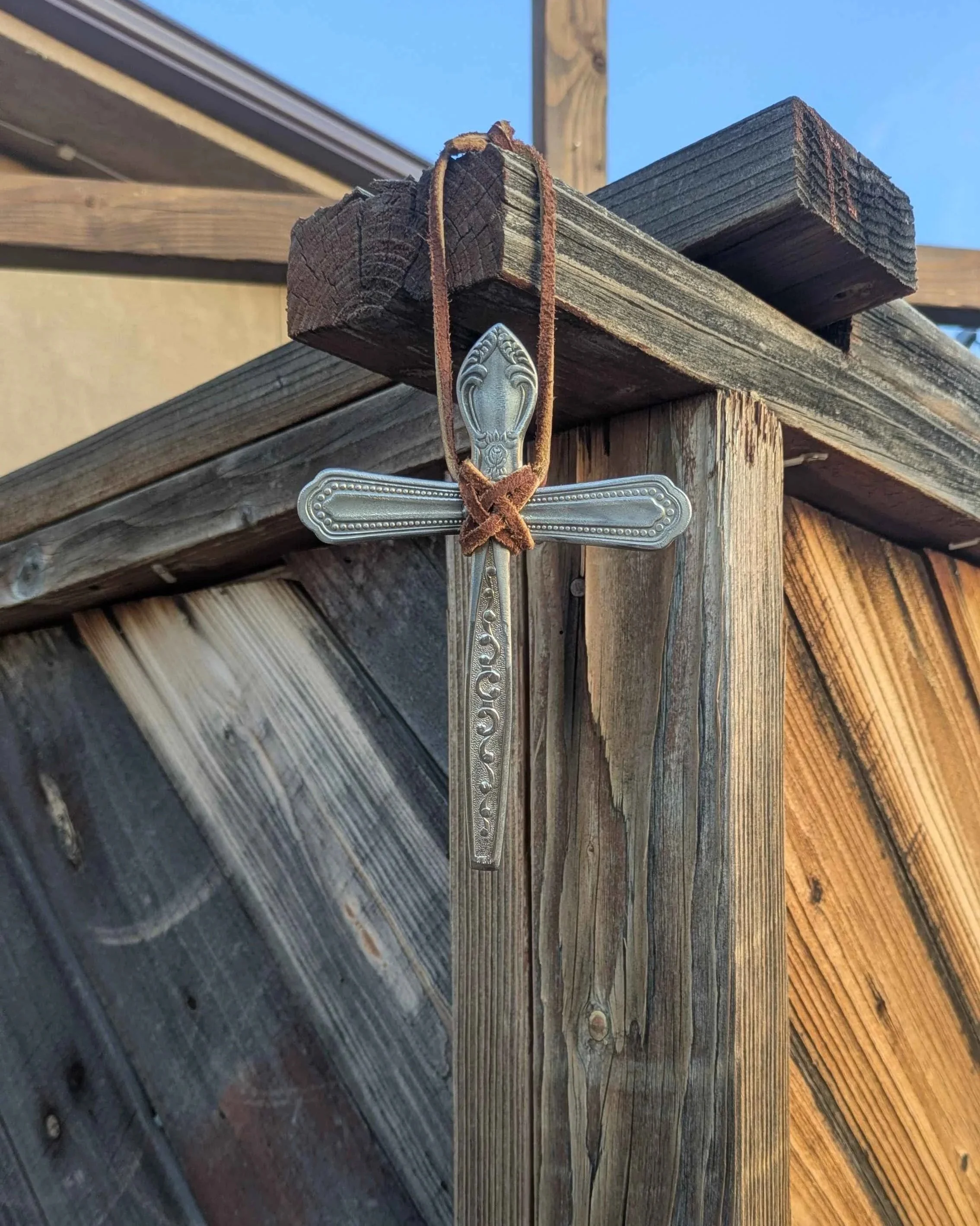 A handmade metal art silverware cross with an ornate leather hanging strap, hung on the side of a fence for home decor purposes..