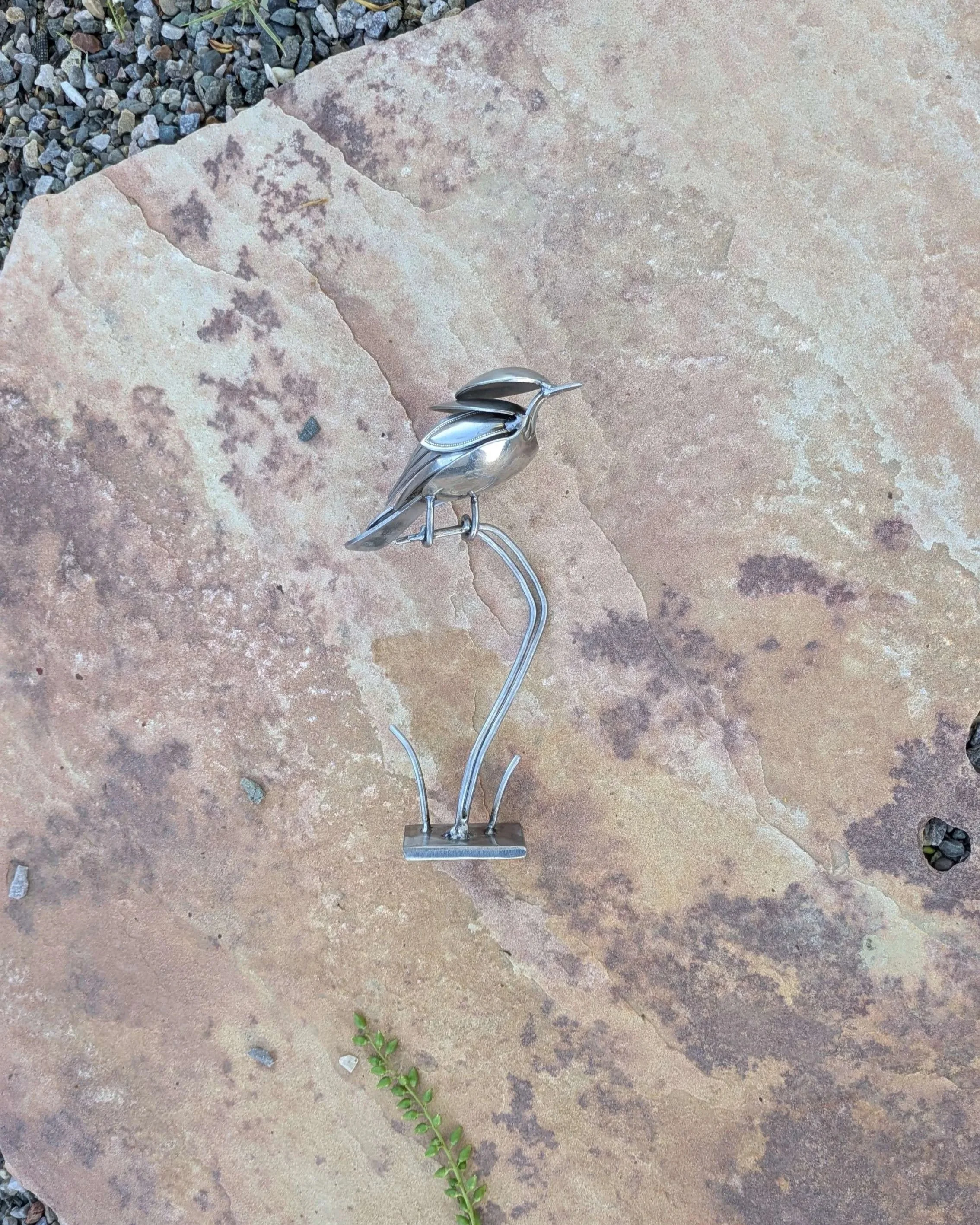 A handmade metal art bird sculpture made from recycled silverware sitting on a wooden shelf, resulting in a great piece of home decor.